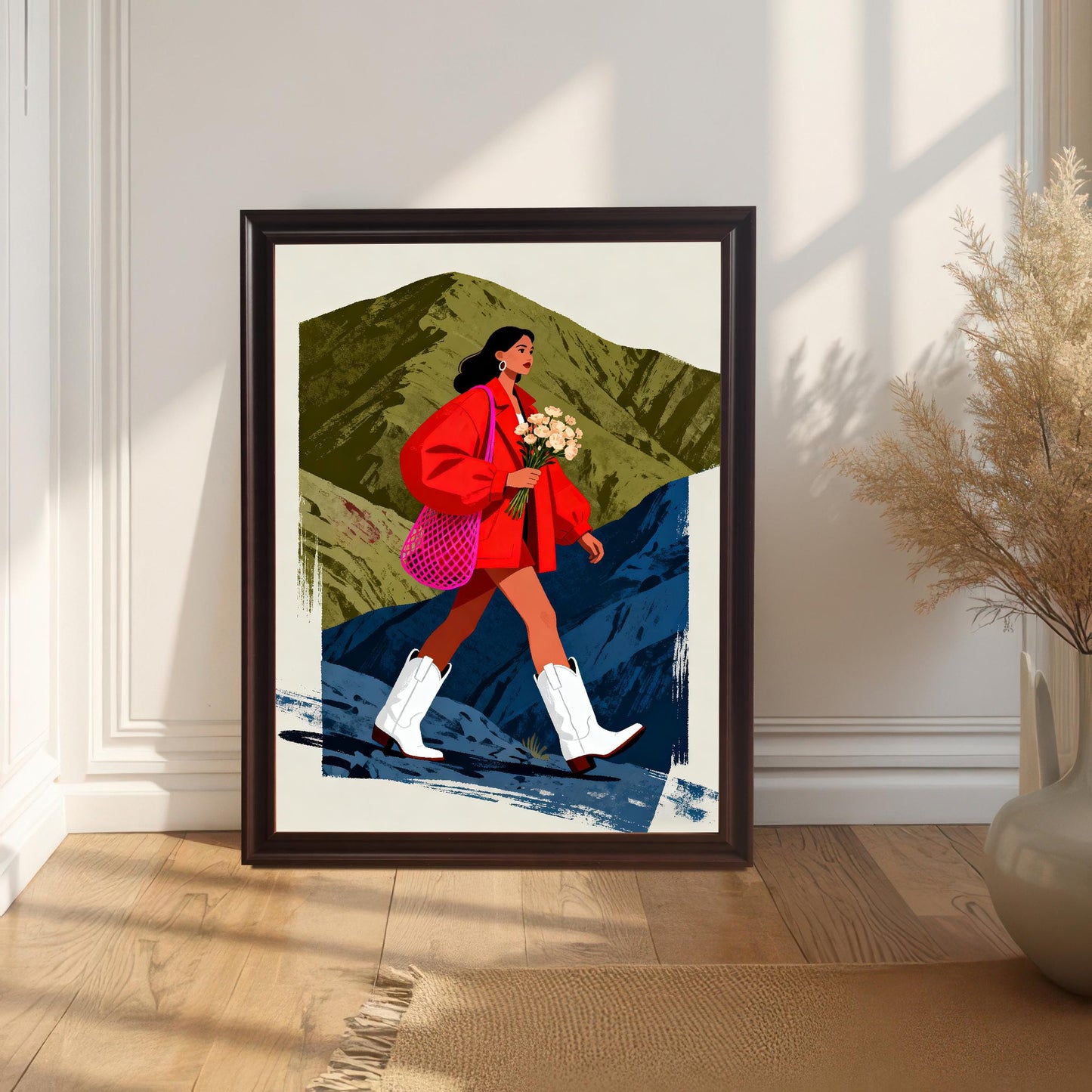 A framed art print featuring a woman in a red jacket and white cowboy boots walking across a stylized mountainous landscape. The print is in a Coffee Frame (dark brown) and is leaning on a hardwood floor against a white, wainscoted wall. A large, dried floral arrangement in a cream vase is on the right.