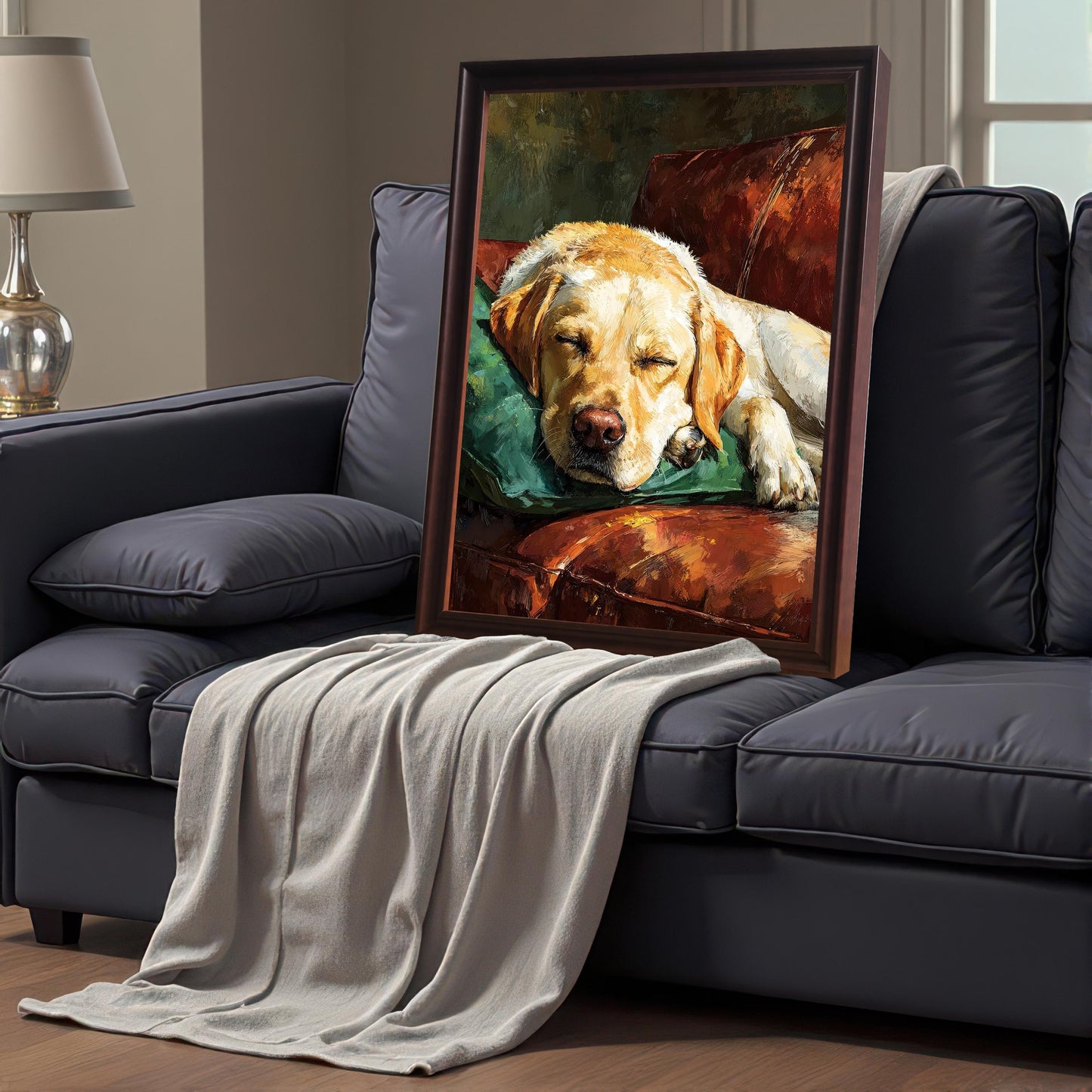 A dark brown-framed painting of a sleeping Labrador on a couch, propped up on a gray sofa draped with a beige blanket.