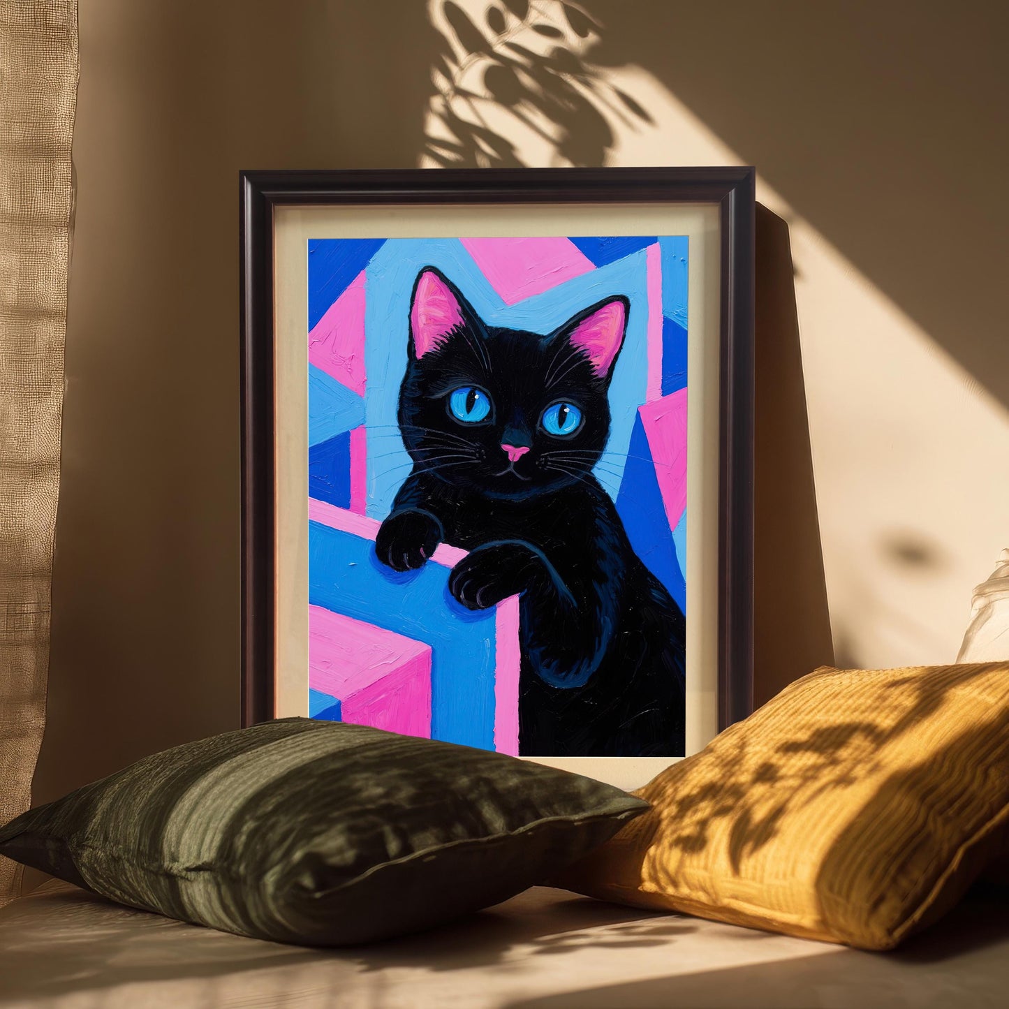 A framed black cat art print in a dark frame, propped up against a wall with pillows in the foreground and a strong sun-drenched shadow.