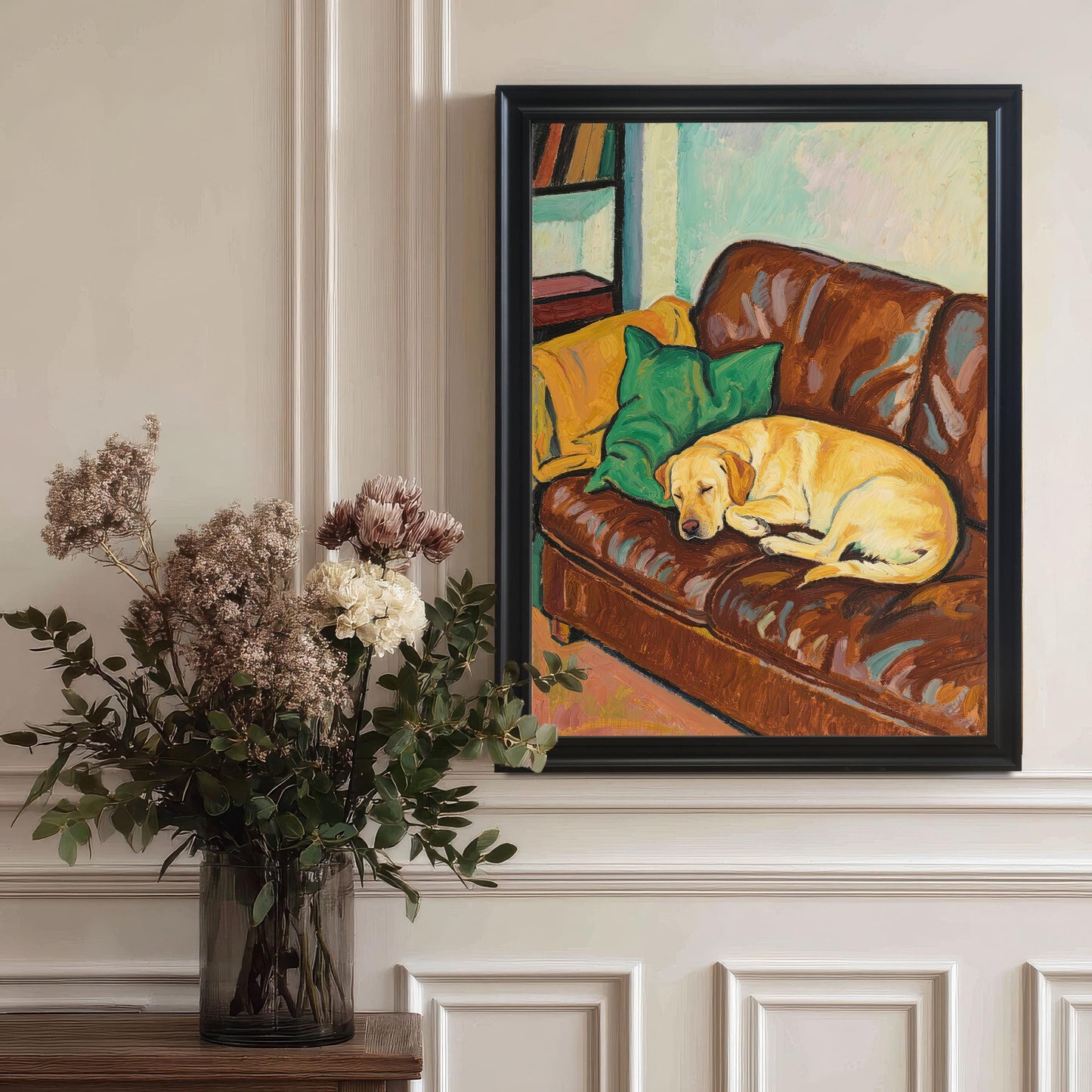 A large, black-framed art print of an oil painting of a yellow Labrador sleeping on a brown leather couch, hanging on a paneled wall near a vase of flowers.