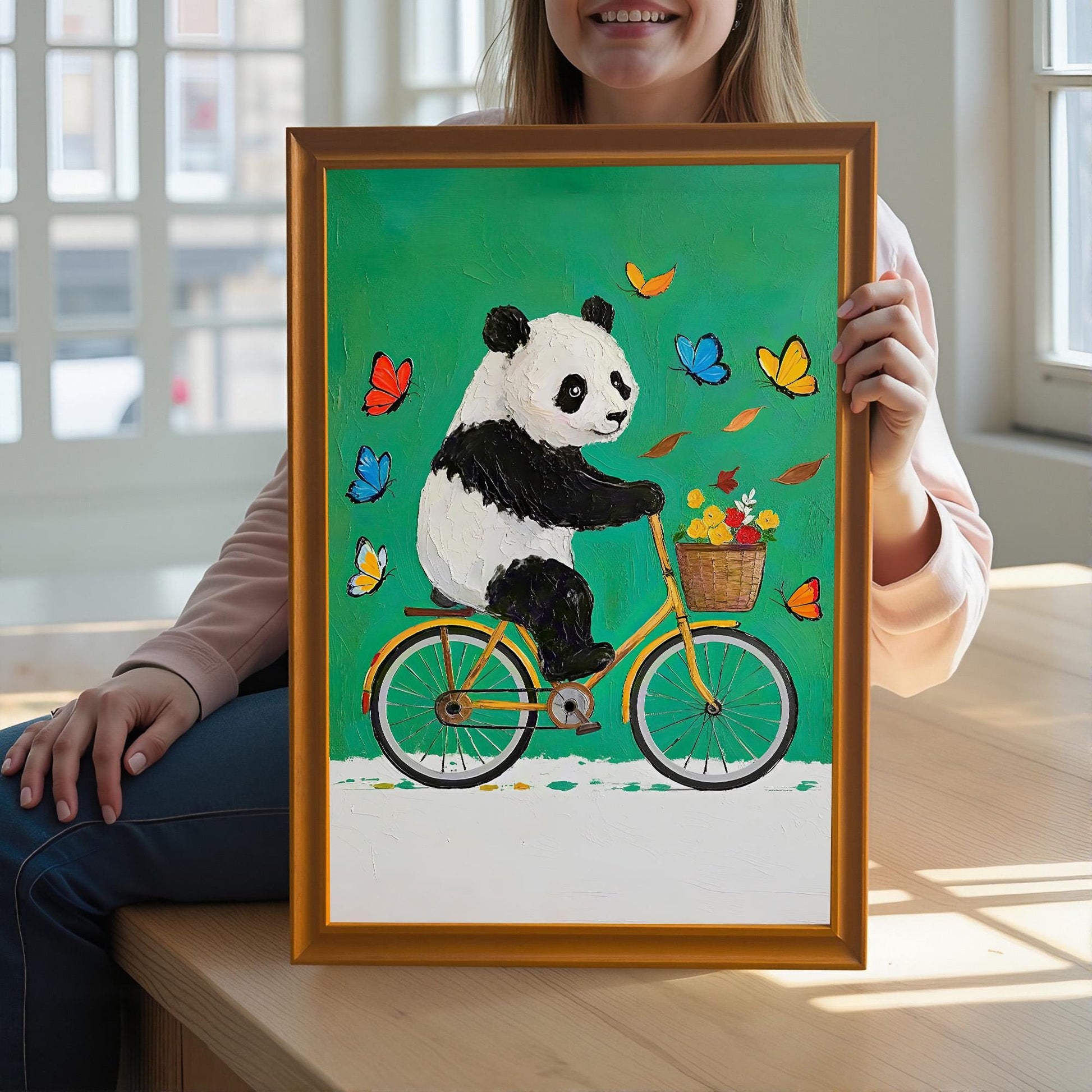 A person holds a framed art print of a panda riding a yellow bicycle with a basket of flowers, framed in a warm brown/orange wood frame.
