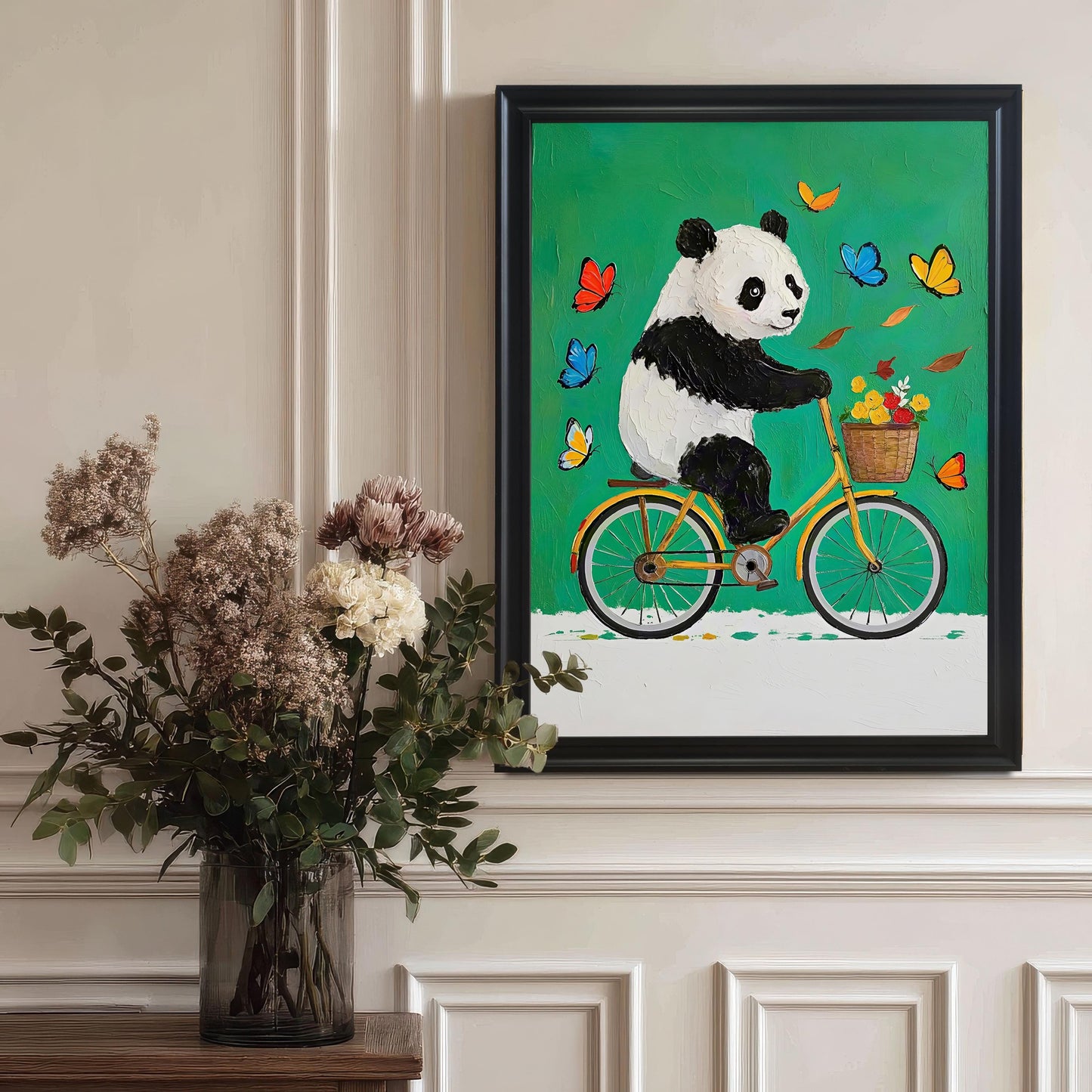 A framed art print of a panda on a bike hangs on a wall next to a large vase of dried flowers. The frame is black.