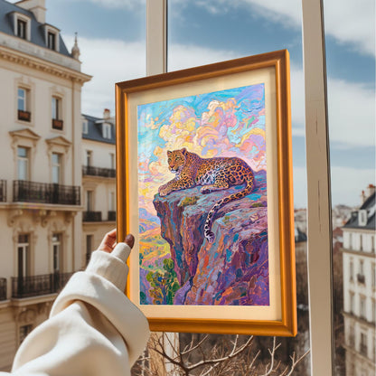 A hand holds up a framed painting of a leopard on a cliff, set against a window with a view of European-style apartment buildings. The frame is orange/gold.
