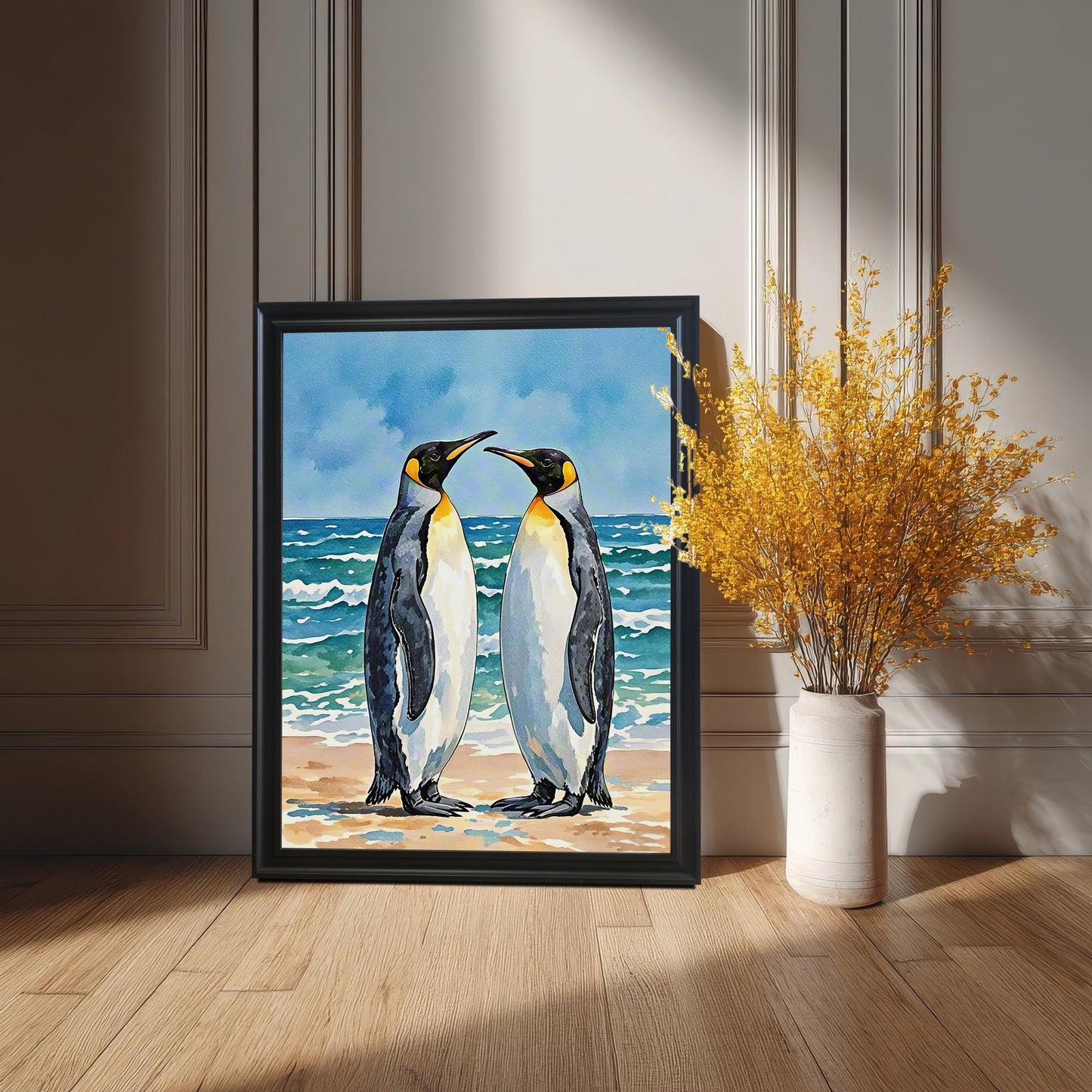 Black-framed watercolor print of two penguins on a beach, leaning on a wooden floor next to a tall vase of yellow dried flowers.