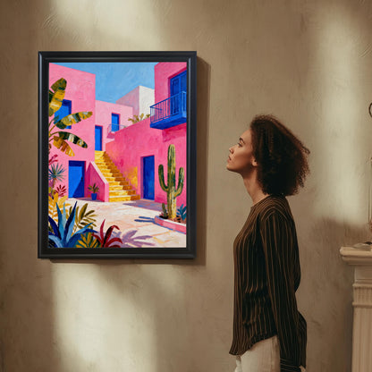A woman with curly hair looks up at a framed painting of a bright pink building with blue accents, yellow stairs, and desert plants (cacti, agave) hanging on a textured wall in a subtly lit room.