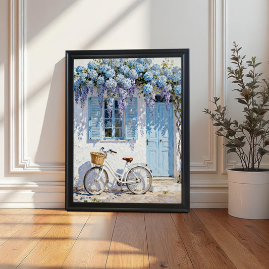 Single framed art print in a Black wood frame leaning against a white wainscoted wall, featuring a charming scene of a white bicycle outside a Greek-style house with blue flowers.