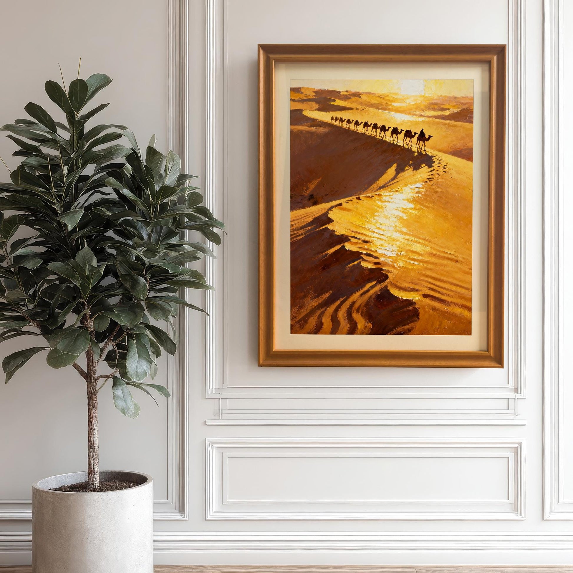 The desert camel caravan art print in a brown/orange wood frame with a mount, hung on a white wainscoting wall next to a large potted fiddle-leaf fig plant, showing the artwork in a home decor setting.