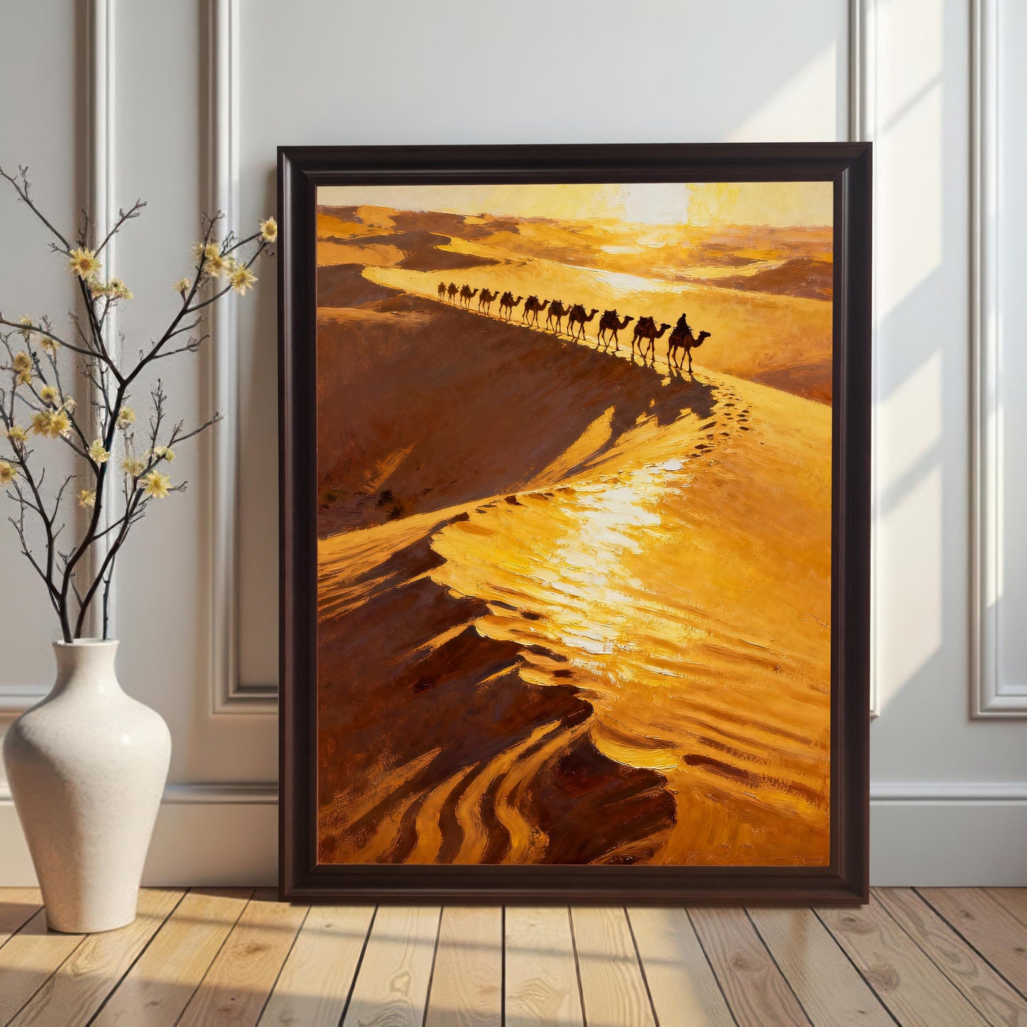 The desert camel caravan art print in a Coffee (dark brown) Frame with No Mount, leaning against a white wainscoting wall on a wooden floor. A white ceramic vase with delicate yellow flowers stands to the left of the frame.