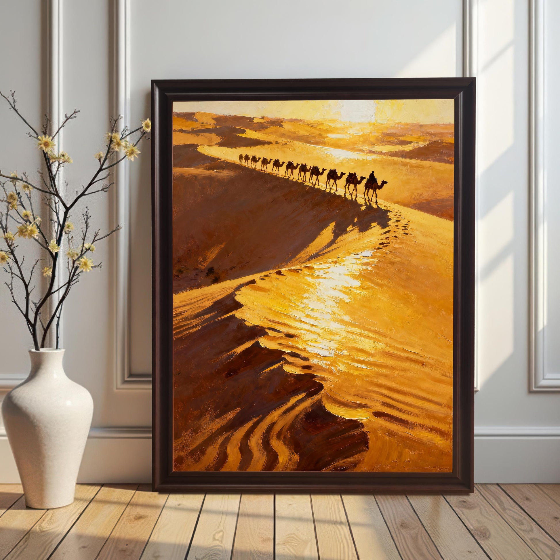 The desert camel caravan art print in a Coffee (dark brown) Frame with No Mount, leaning against a white wainscoting wall on a wooden floor. A white ceramic vase with delicate yellow flowers stands to the left of the frame.