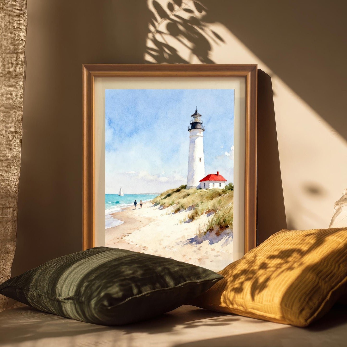 Close-up lifestyle photo showing the framed art print of a lighthouse on a beach leaning against a wall, surrounded by pillows in a warmly lit, casual setting. The frame is a natural wood color.