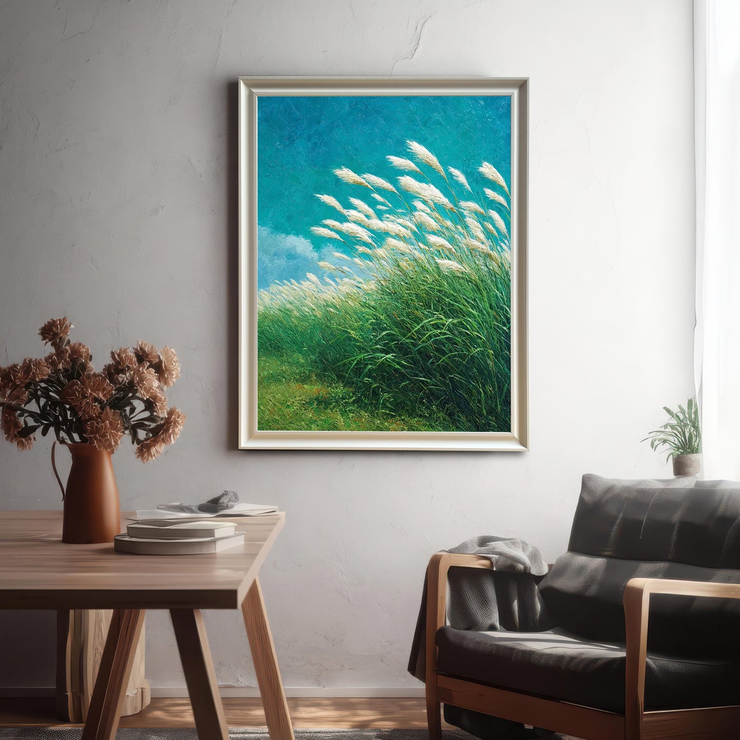Lifestyle photo showing a framed art print of white, feathery grass blowing in a green field under a bright blue sky. The print is hung above a wooden table and a dark armchair in a modern living space. The frame is a light, possibly silver or natural wood, color.