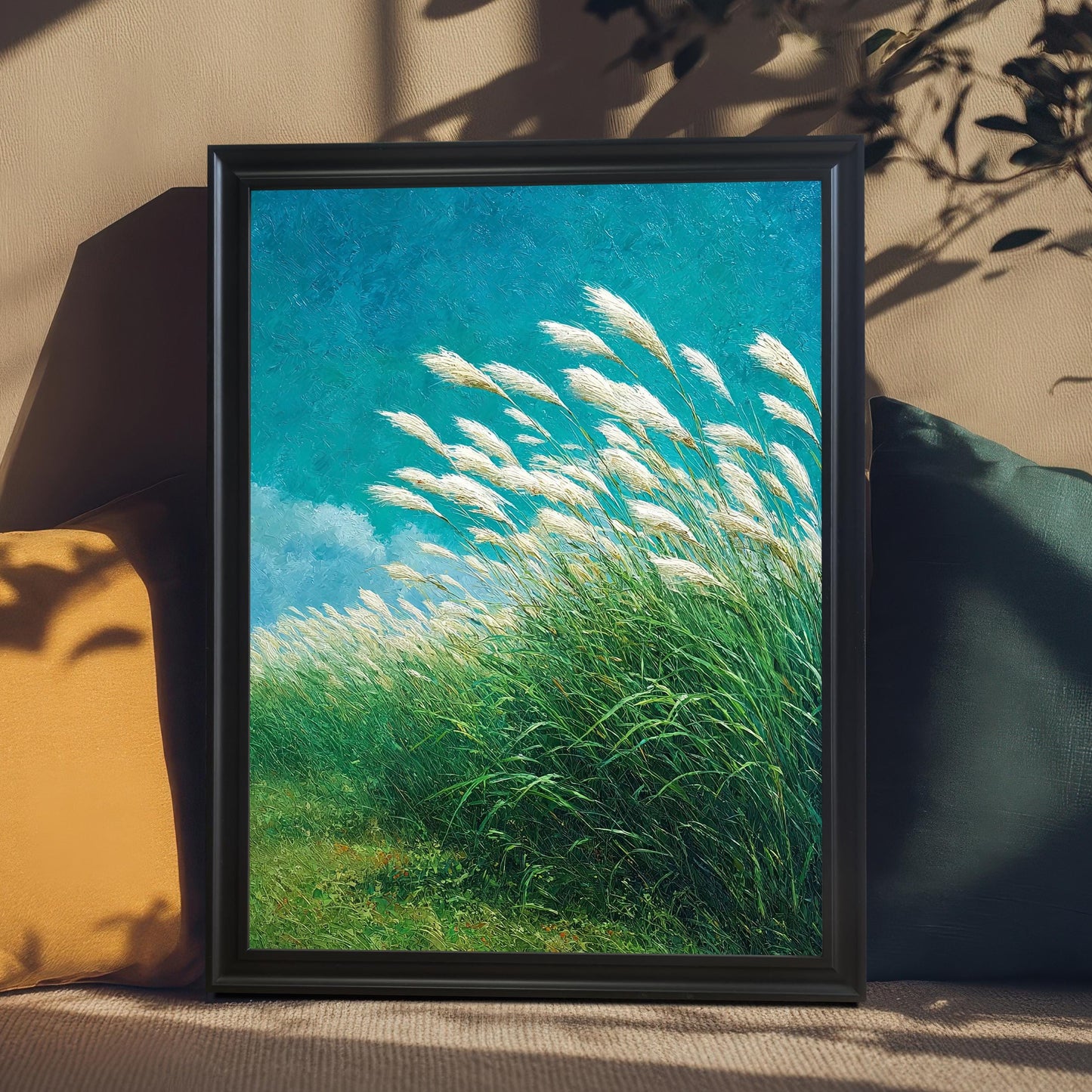 Black wood-framed art print of a textured painting showing a field of green grass and white feathery reeds (pampas grass) under a striking turquoise sky. The print is styled leaning on a beige couch with a yellow pillow and a dark teal pillow, with sunlight and plant shadows casting across it.