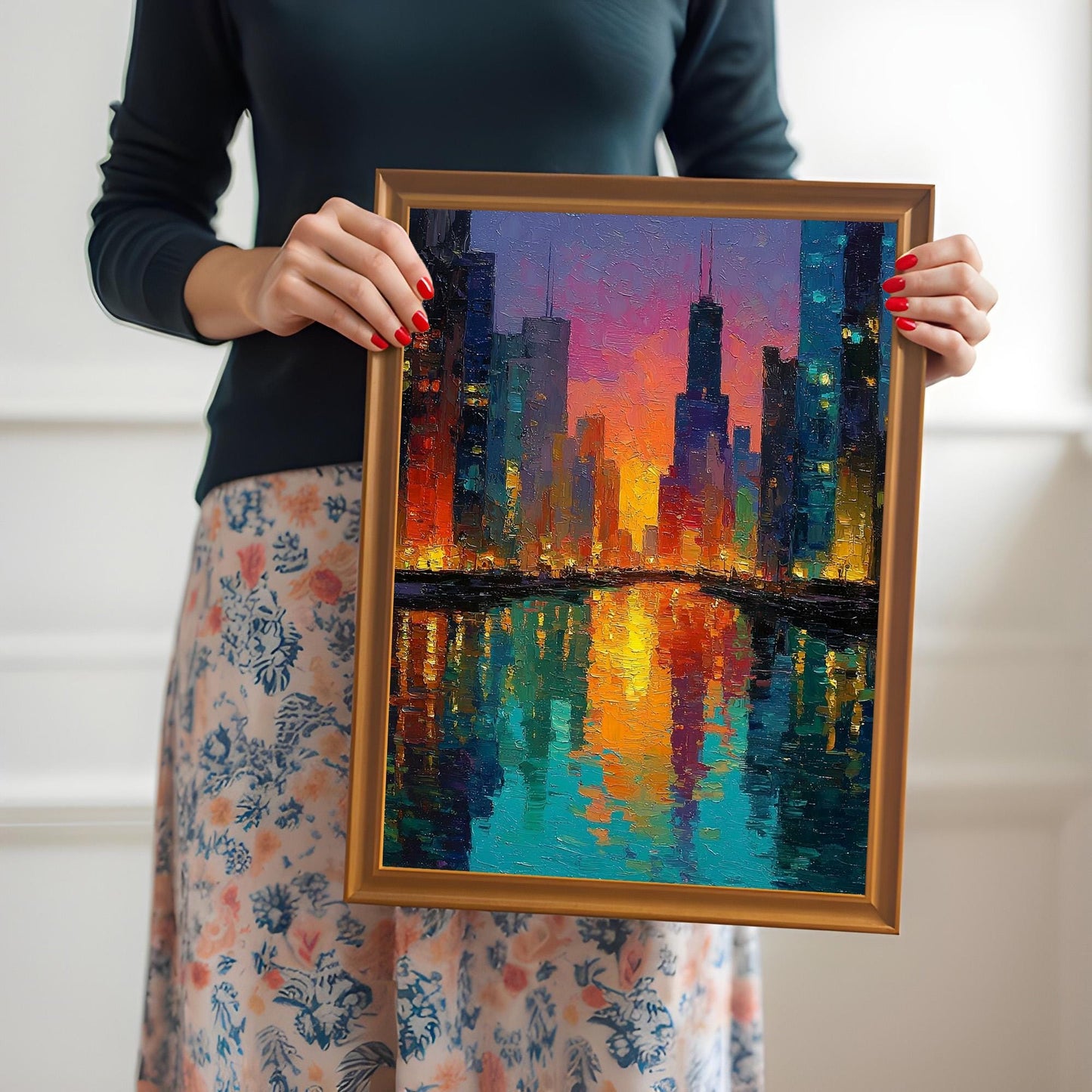A person wearing a dark top and a floral skirt holds up a gold-framed print of a colorful, impressionist cityscape painting (Chicago skyline) with vibrant reflections. The print is held near the chest against a light, minimalist background.