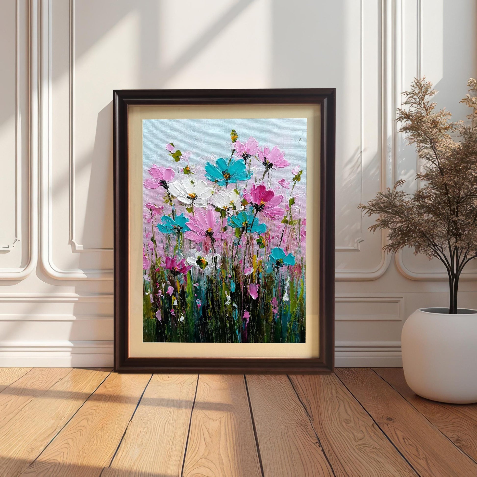 Dark brown wood-framed art print of a textured floral oil painting with pink, turquoise, and white flowers in a field of green. The print is leaning on a light wood floor against a decorative white paneled wall with shadows from a window, next to a tall white planter.