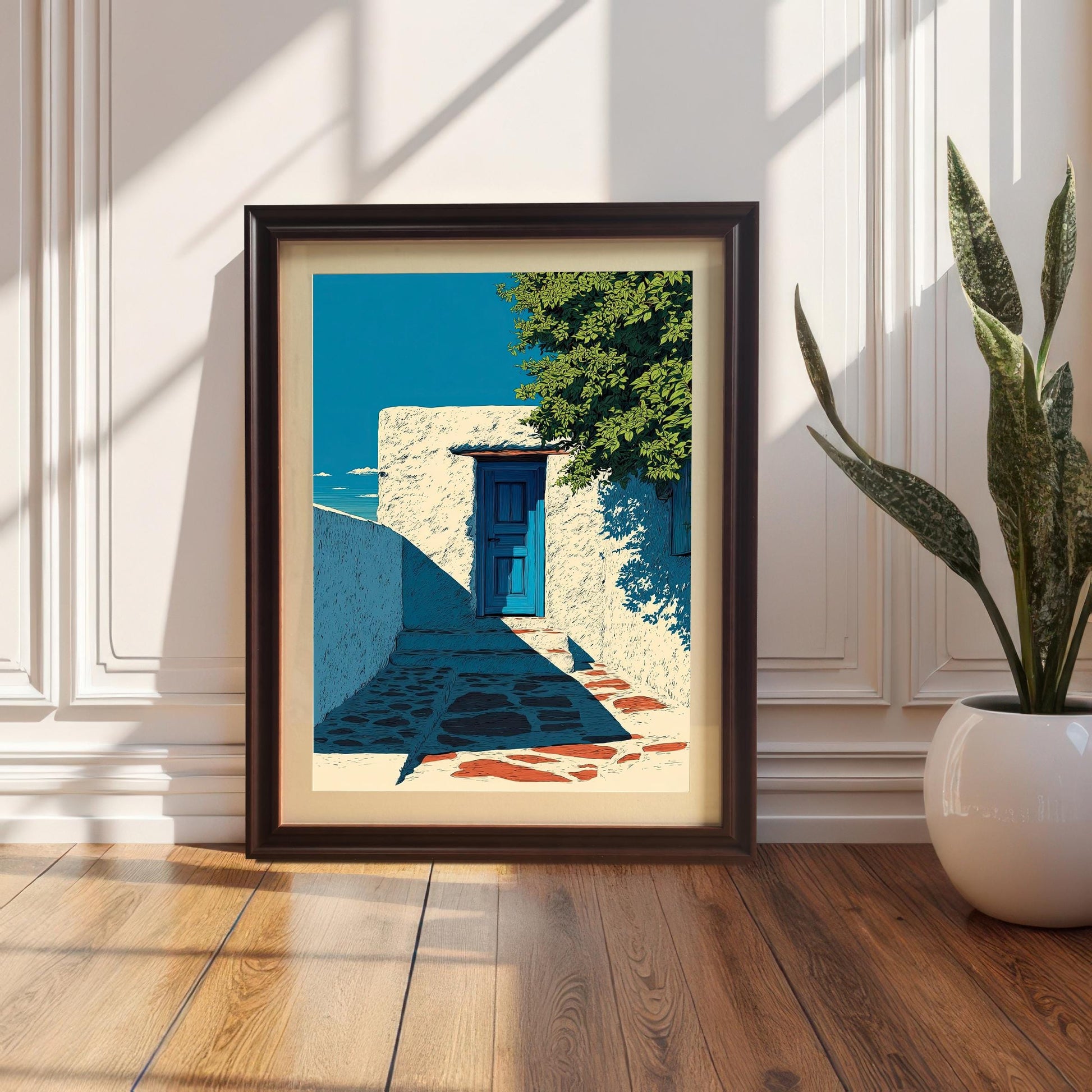 The art print of a white stucco wall and blue door, displayed in a Coffee Wood Frame with a Mount (matting). The frame is leaning on a wood floor against a white wainscoted wall next to a potted snake plant, with bright sunlight shining.