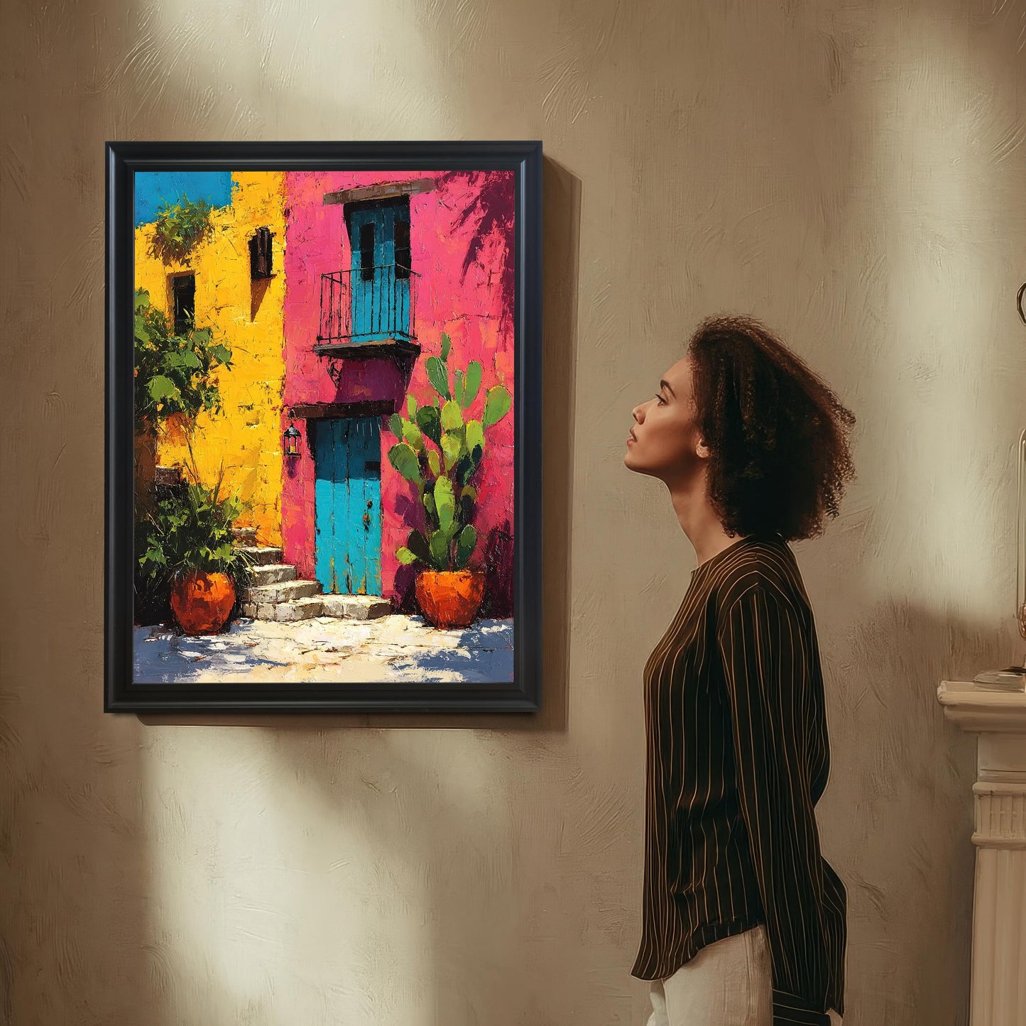 A woman with curly hair, wearing a striped shirt, stands looking up at the framed art print of a bright, textured building in yellow, pink, and blue, which is hanging on a textured beige wall. The frame is Black wood.