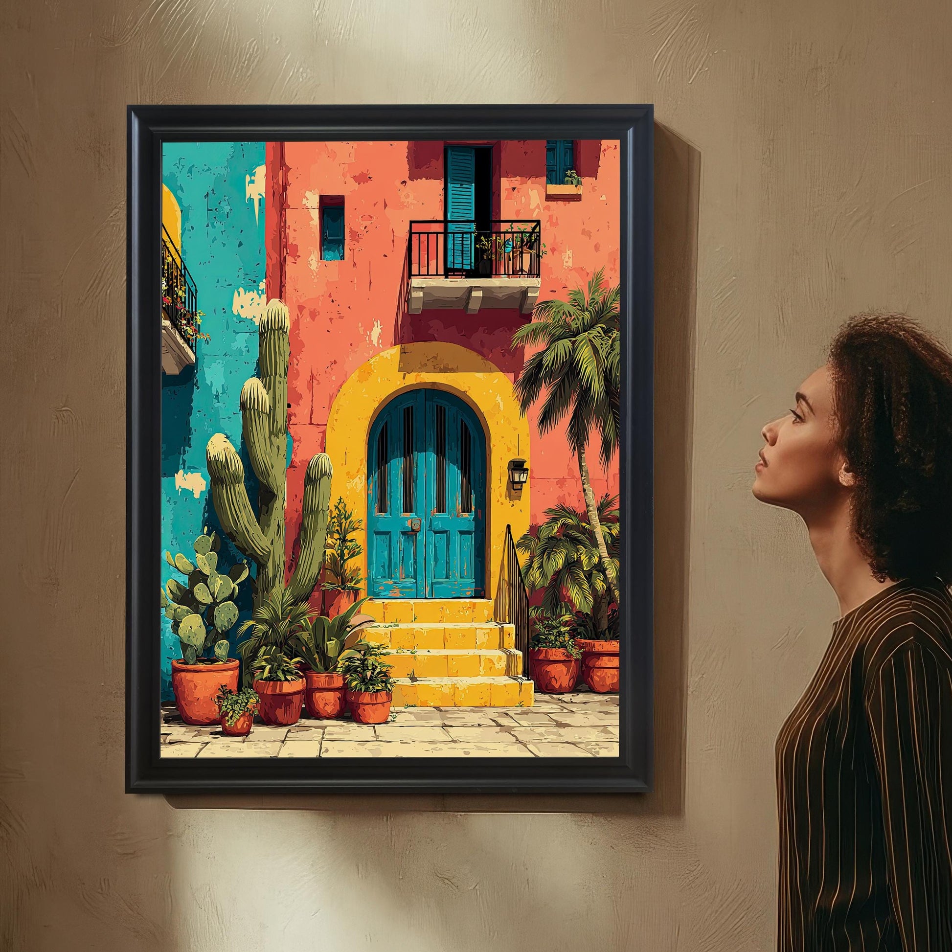 A woman with curly hair, wearing a striped shirt, views the framed art print (in a sleek Black Frame without a mount/mat) hanging on a textured wall. The art depicts a vibrant Mexican-style house with a yellow arch, blue door, and cacti.