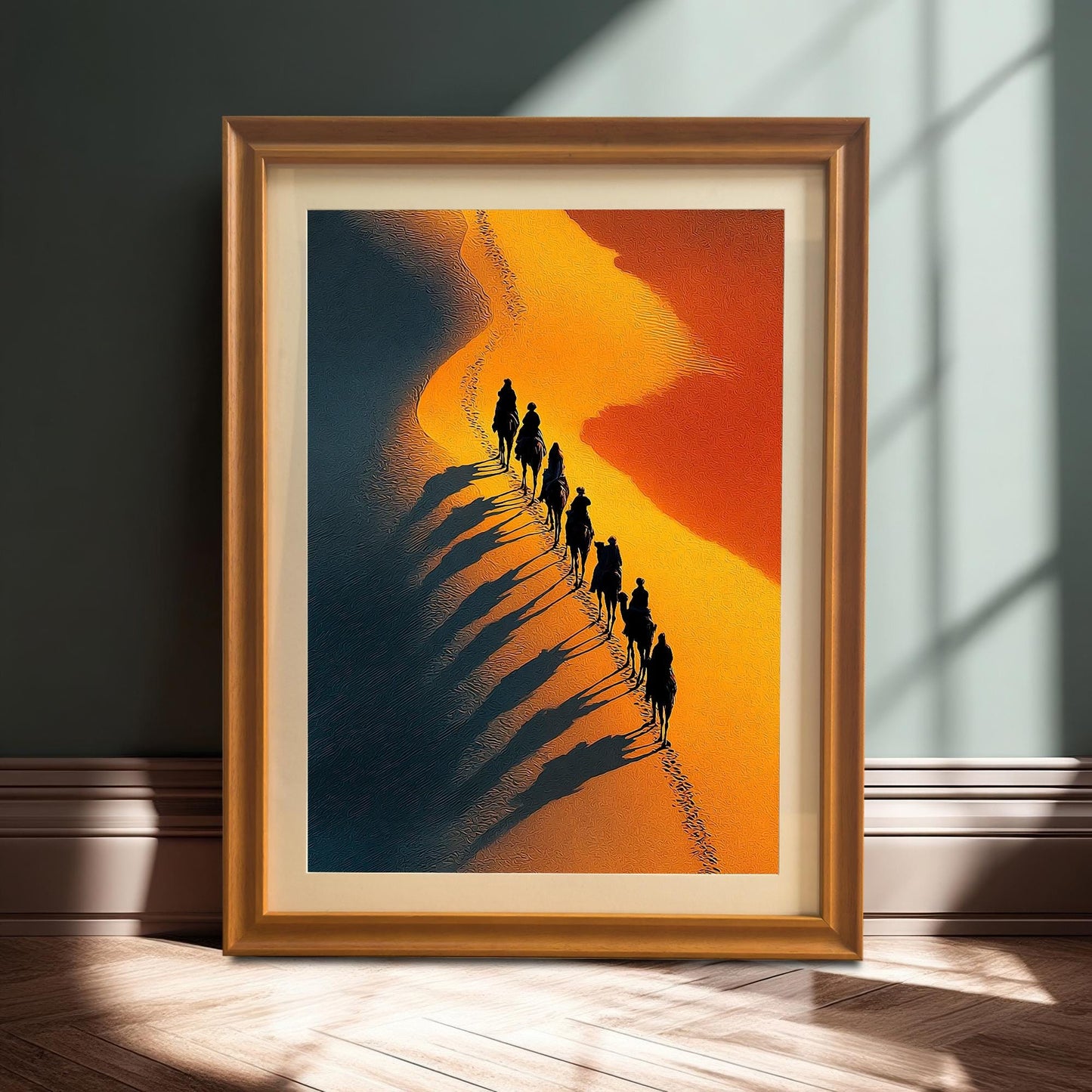 Stylized photo of the framed desert art print (Orange Frame & Mount) resting on a wooden floor against a dark wall, illuminated by a strong sunlight shadow. The art shows a line of camels with long shadows on a sandy dune.