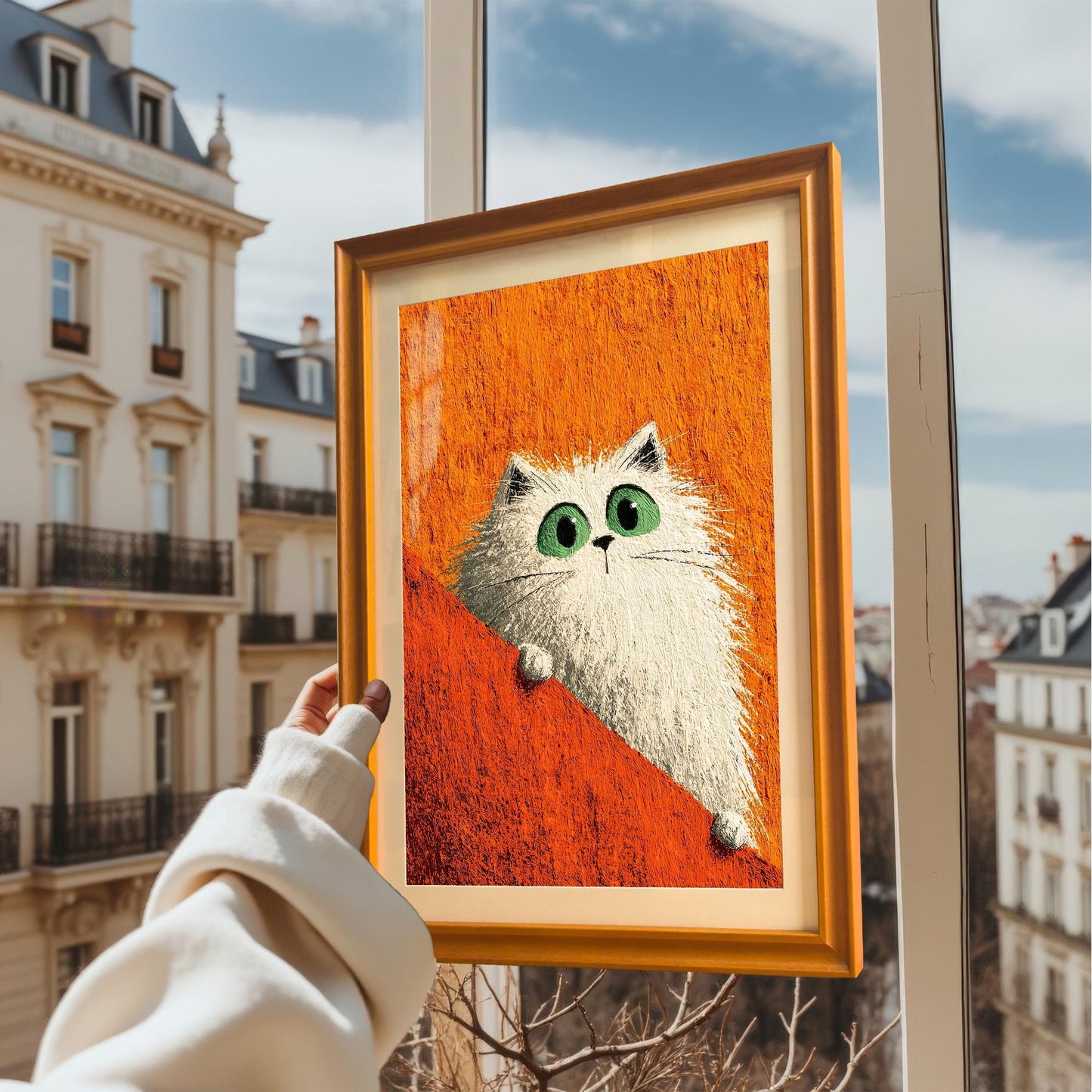 A hand in a cream sweater holds up a framed art print of a fluffy white cat with big green eyes against an orange background, viewed through a window with a European city street outside.