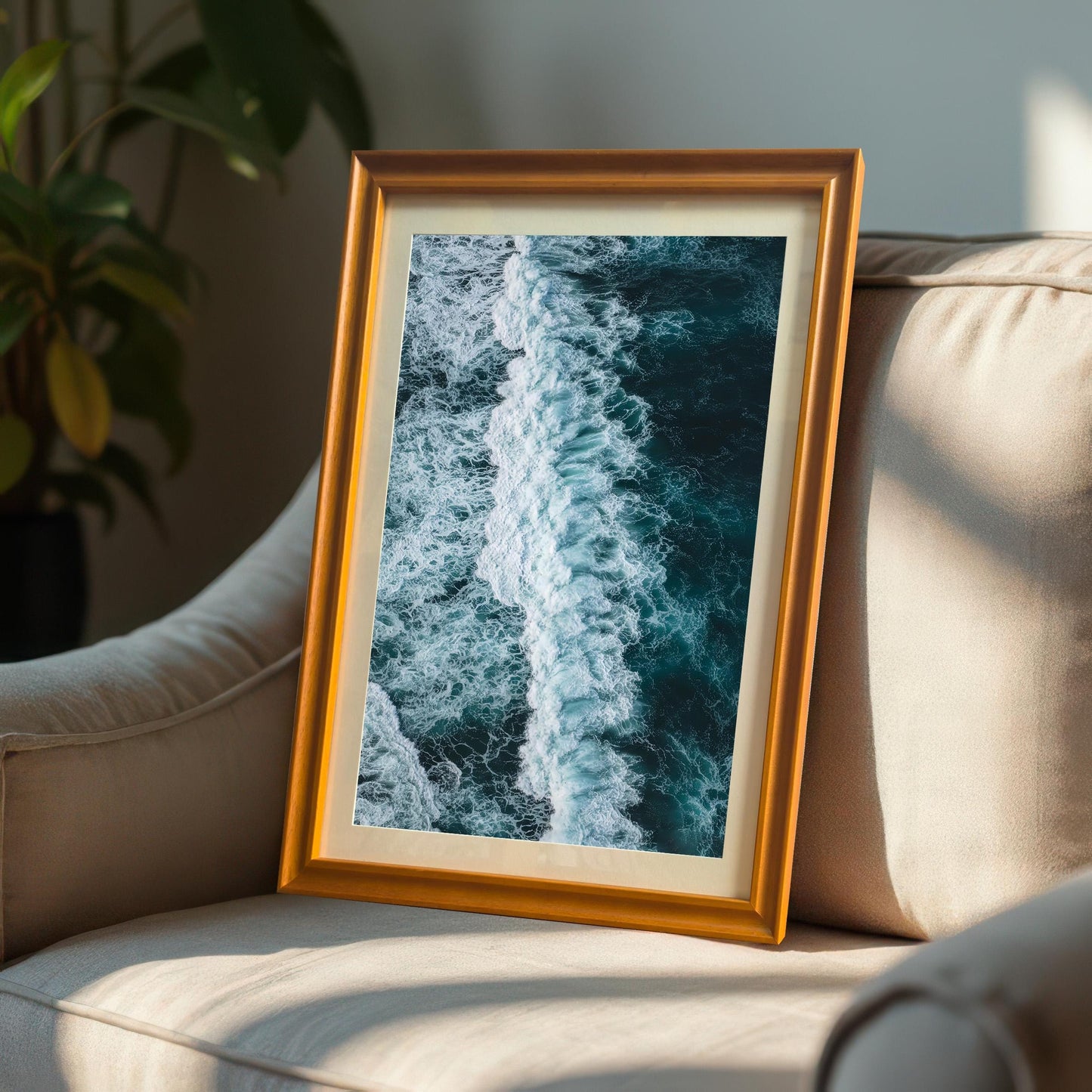 Vertical, portrait-oriented framed print of an ocean wave, with an Orange/Gold frame and a mount, leaning casually against a light-colored armchair in a sunny room.