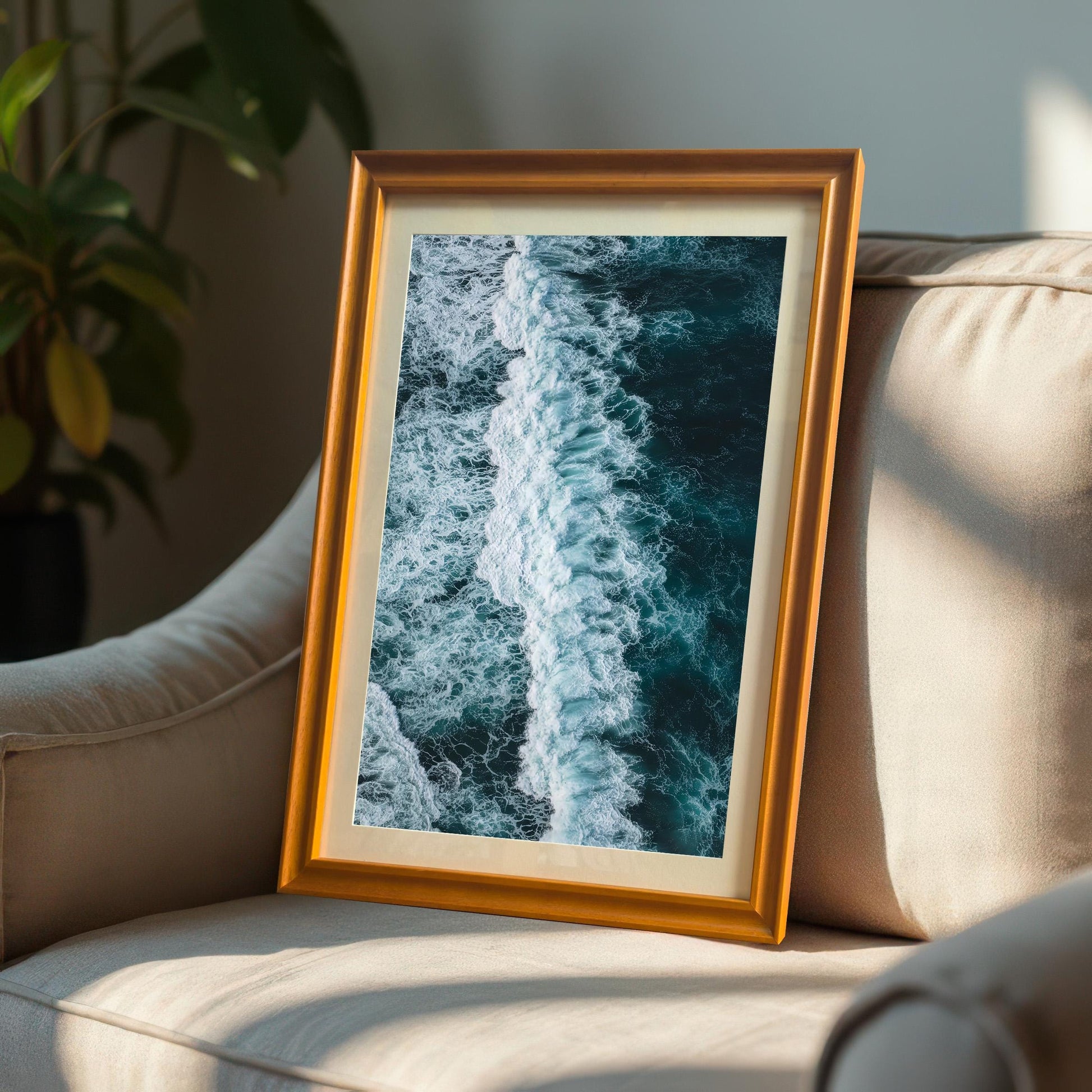 Vertical, portrait-oriented framed print of an ocean wave, with an Orange/Gold frame and a mount, leaning casually against a light-colored armchair in a sunny room.