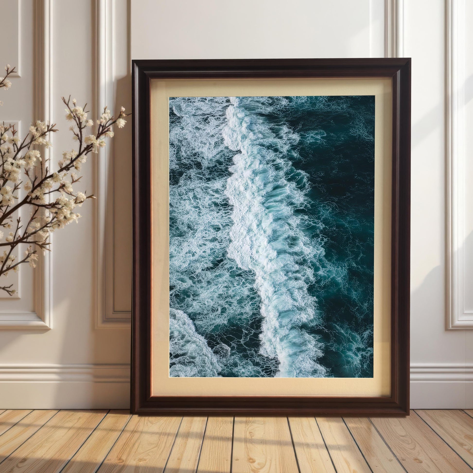 Vertical, portrait-oriented framed print of an ocean wave, with a Coffee brown wood frame and a mount (matting), leaning on a wooden floor against a white paneled wall next to a branch of white flowers.