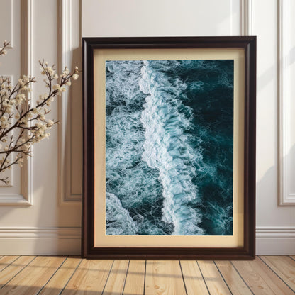 Vertical, portrait-oriented framed print of an ocean wave, with a Coffee brown wood frame and a mount (matting), leaning on a wooden floor against a white paneled wall next to a branch of white flowers.