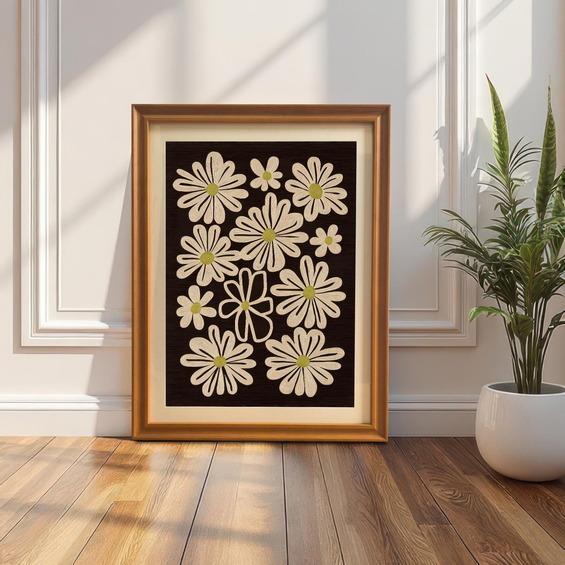 Orange framed art print of retro daisy flowers, leaning on the floor against a white paneled wall next to a potted snake plant.