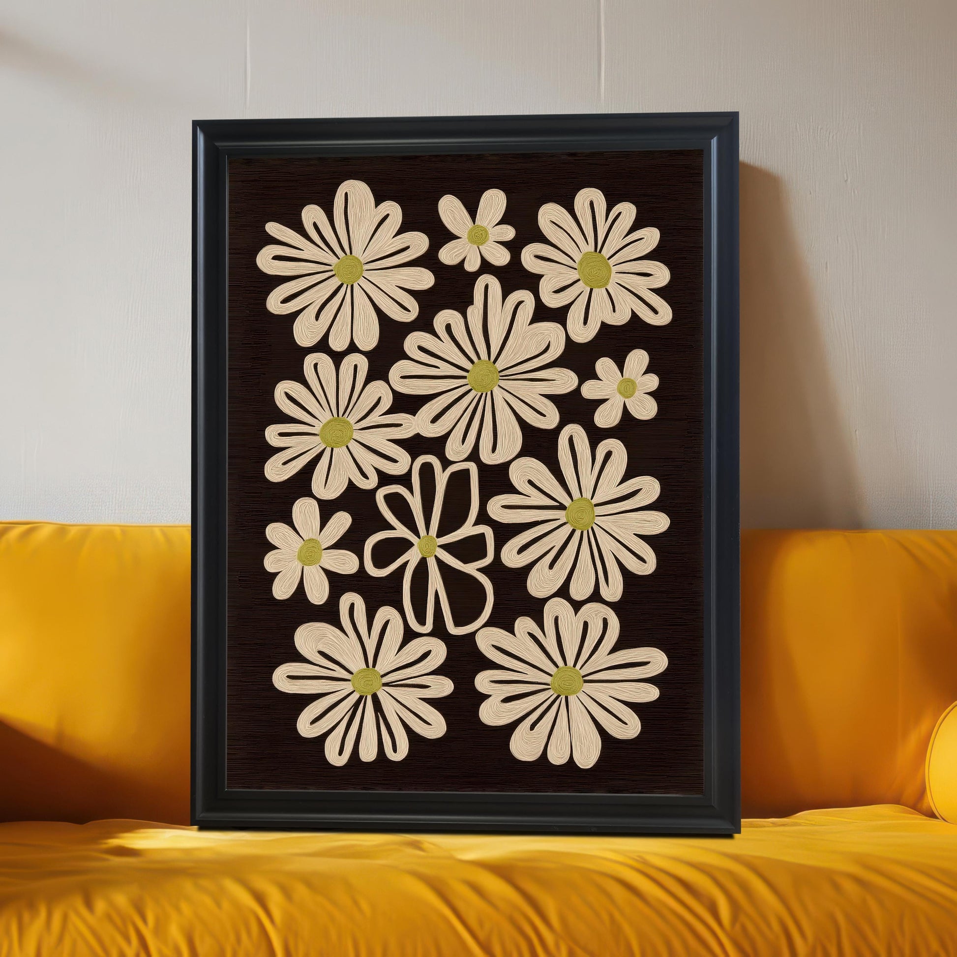 Black framed art print of retro daisy flowers on a dark background, leaning against a wall and resting on a bright yellow sofa.