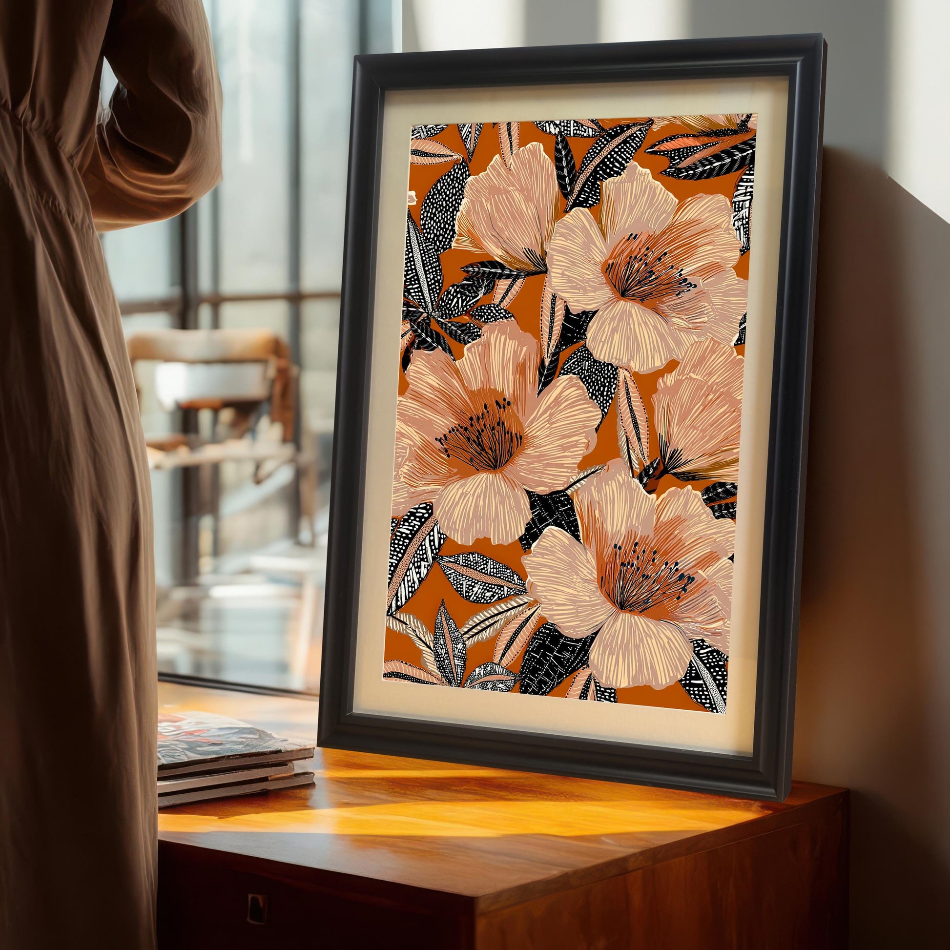 Lifestyle photo of the floral art print in a Black wood frame with a mat/mount, standing on a wooden cabinet near a large window, next to a person in a brown dress. The artwork is a maximalist design with large peach and pink floral elements on a warm orange backdrop.