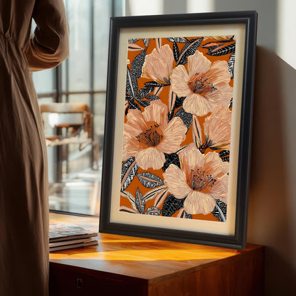 Lifestyle photo of the floral art print in a Black wood frame with a mat/mount, standing on a wooden cabinet near a large window, next to a person in a brown dress. The artwork is a maximalist design with large peach and pink floral elements on a warm orange backdrop.