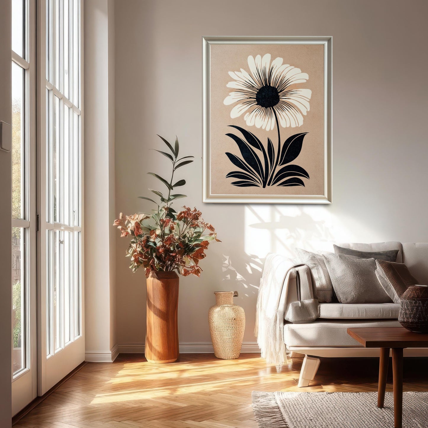 Lifestyle photo of the framed flower art hanging on a light wall in a bright living room. The print is in a White wood frame and features a graphic white flower with black details on a beige backdrop, placed above a neutral couch and tall floor vases.