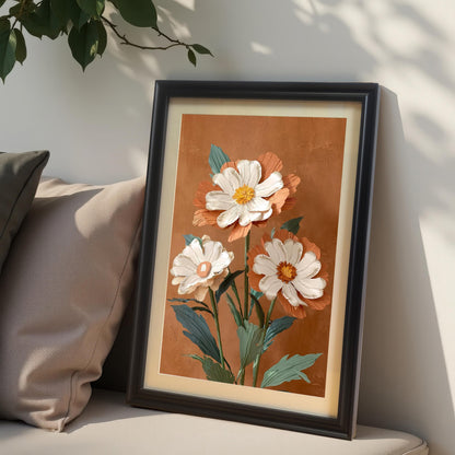Close-up lifestyle photo of the floral art print in a Black wood frame with a mat/mount, leaning against a neutral wall with beige pillows. The print showcases an impasto-style artwork of three white and burnt-orange flowers.