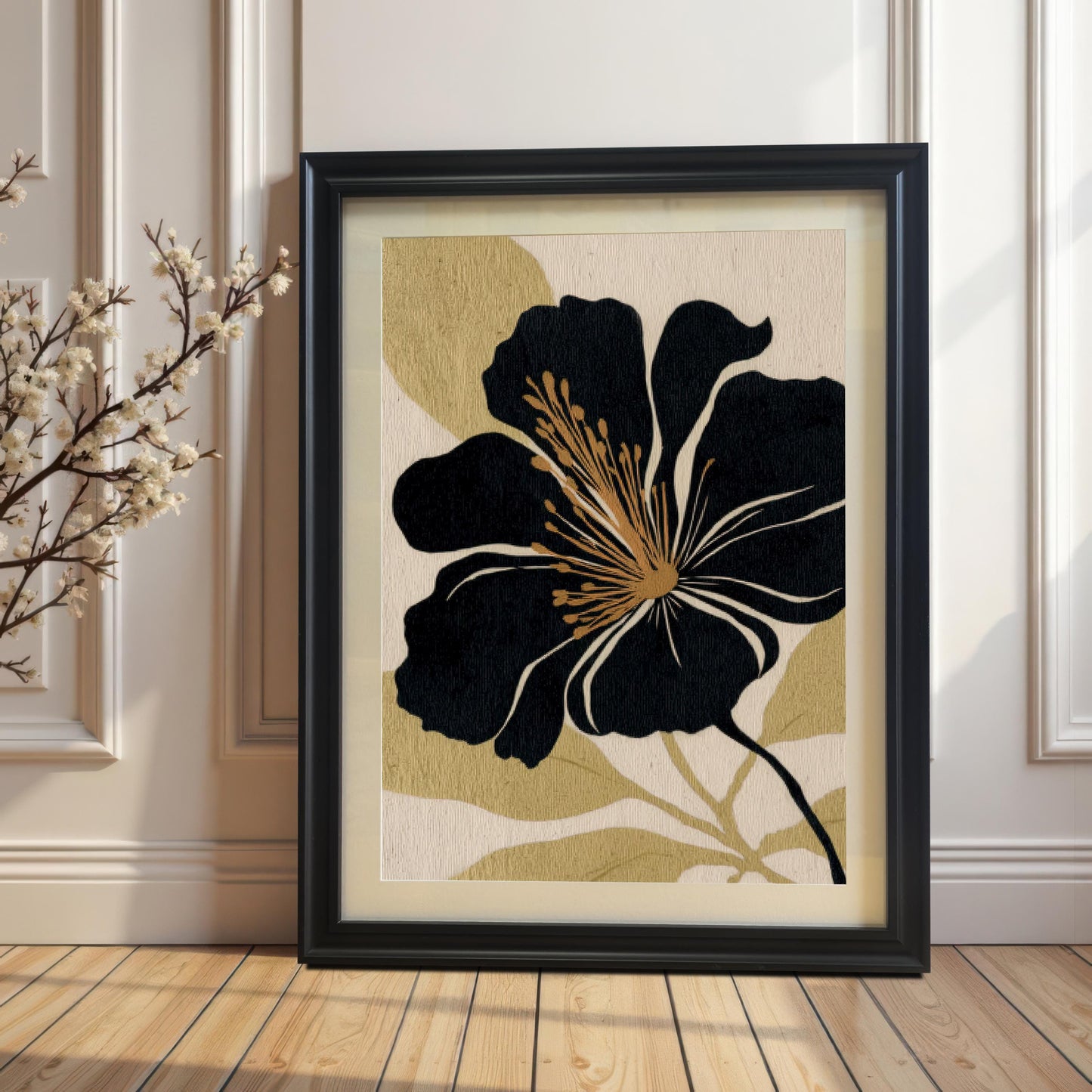 The black and gold abstract floral art print in a Black Frame with a Mount (matting) rests on a wooden floor against a white paneled wall. A branch with small white blossoms is visible on the left, highlighting the contrast and natural aesthetic.