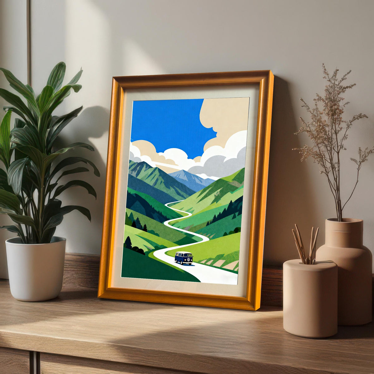 The framed mountain road trip print in a warm Orange/Gold Frame leans on a wooden surface next to a potted plant, showcasing the artwork's colors in a bright setting.