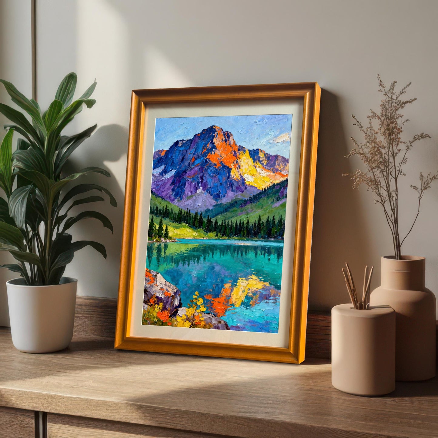 Vibrant mountain and lake art print in a gold/orange frame with a mount leaning on a wooden shelf next to a potted plant and ceramic vases.