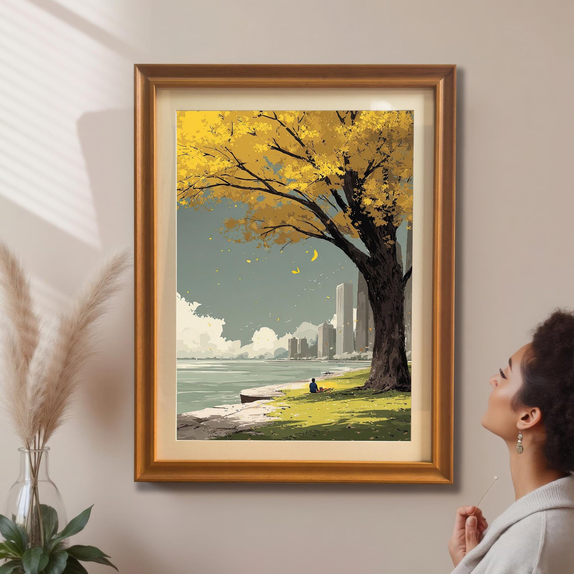A woman admires the yellow autumn tree and city park art print which is framed in a gold/orange frame with a mount and hanging on a beige wall.