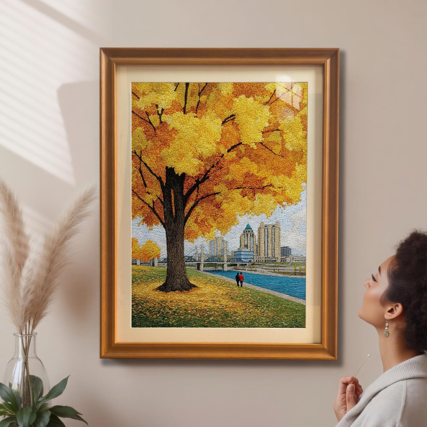 A woman admires the textured yellow autumn tree and cityscape art print which is framed in a gold/orange frame with a mount and hanging on a beige wall.