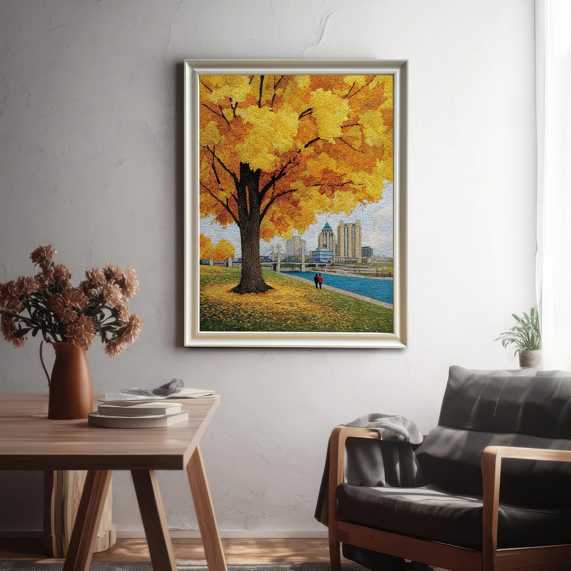 Textured yellow autumn tree and cityscape art print in a white/silver frame with a mat/mount. The framed artwork is hanging on a white wall in a living space, above an armchair and next to a wooden table with a vase.