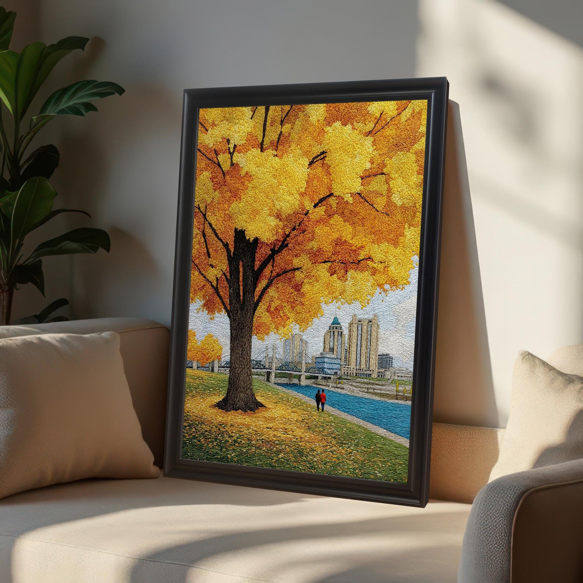 Textured yellow autumn tree and cityscape art print with a couple walking by the river. The artwork is shown in a black wood frame leaning on a neutral-colored couch in a sunny living room setting.