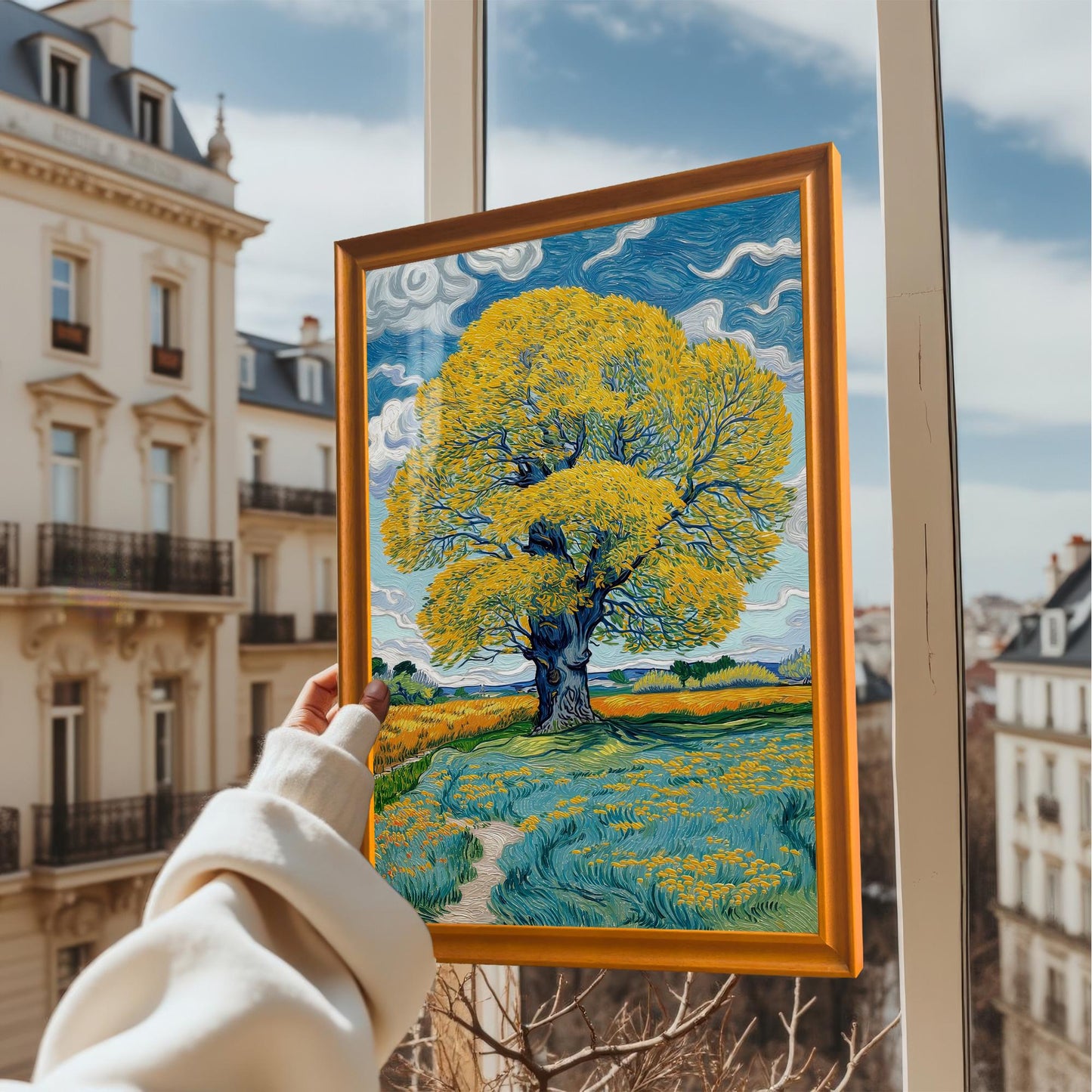 A person holds the framed art print with an Orange frame up to a window overlooking a European cityscape. The print depicts a vibrant yellow tree against a blue, cloudy sky.