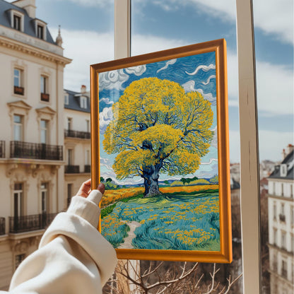 A person holds the framed art print with an Orange frame up to a window overlooking a European cityscape. The print depicts a vibrant yellow tree against a blue, cloudy sky.