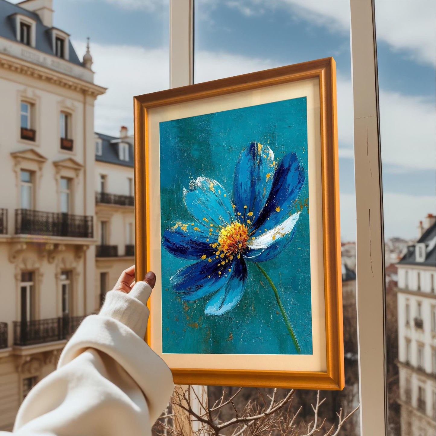 A person holds the framed art print with an Orange frame up to a window overlooking a European cityscape. The print depicts a vibrant blue and gold flower.