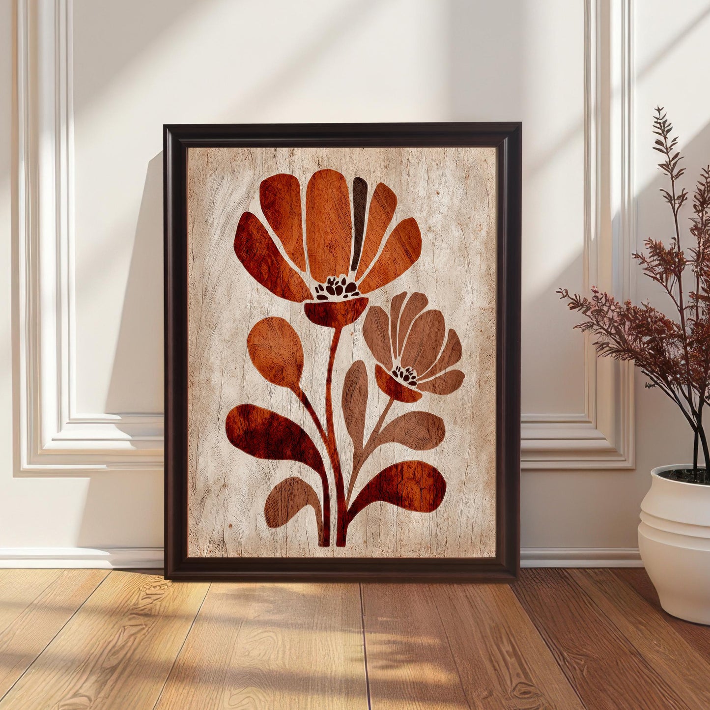 Vertical framed art print of two stylized, modern, rust-orange and brown flowers on a light, distressed wood-texture background, framed with a black frame. The print is leaning on a hardwood floor against a white wall with wainscoting, next to a white potted plant. (This describes the setting and frame color.)