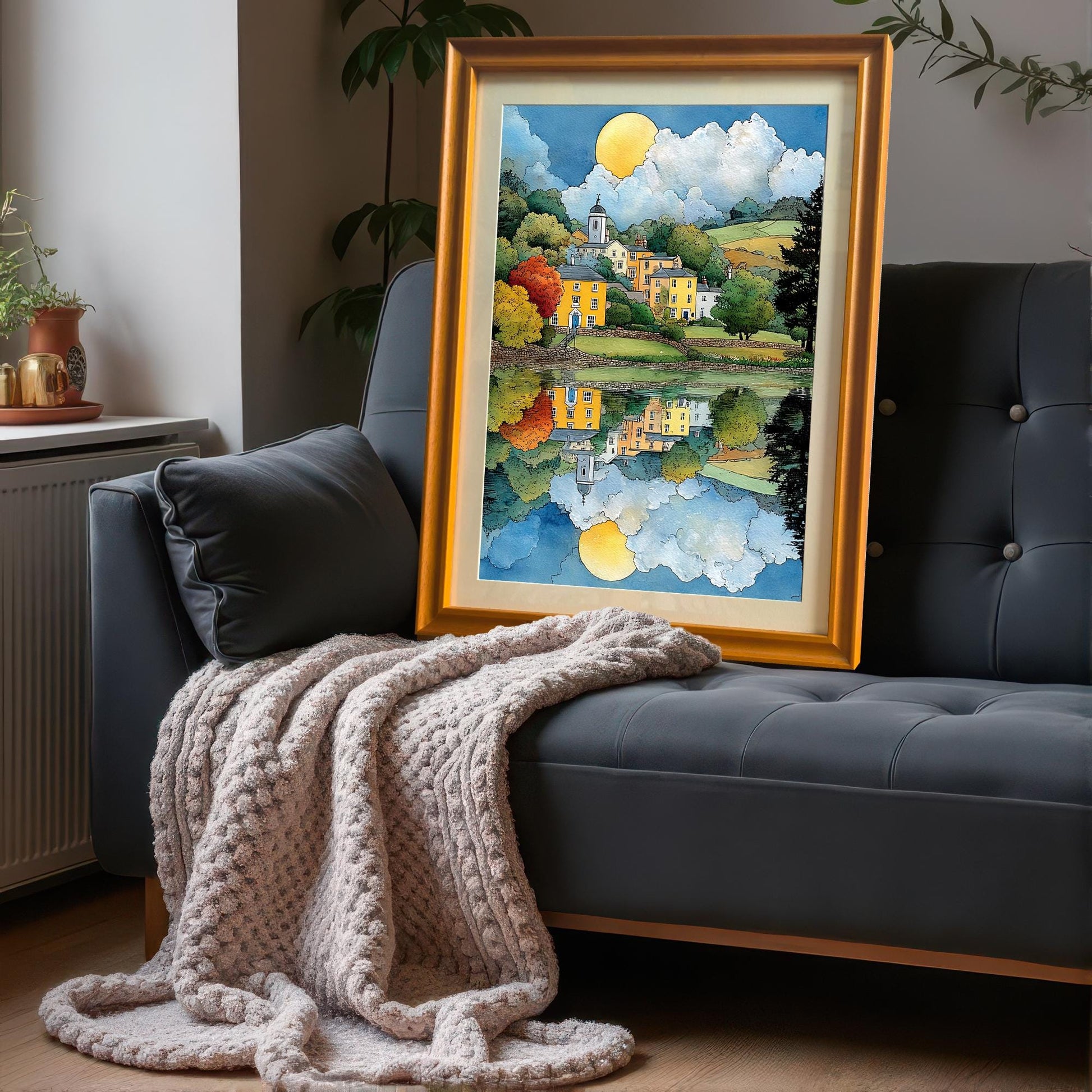 Framed art print leaning on a dark grey sofa with a knitted throw blanket. The frame is a bold orange or gold color. The art depicts a brightly colored village and its reflection.