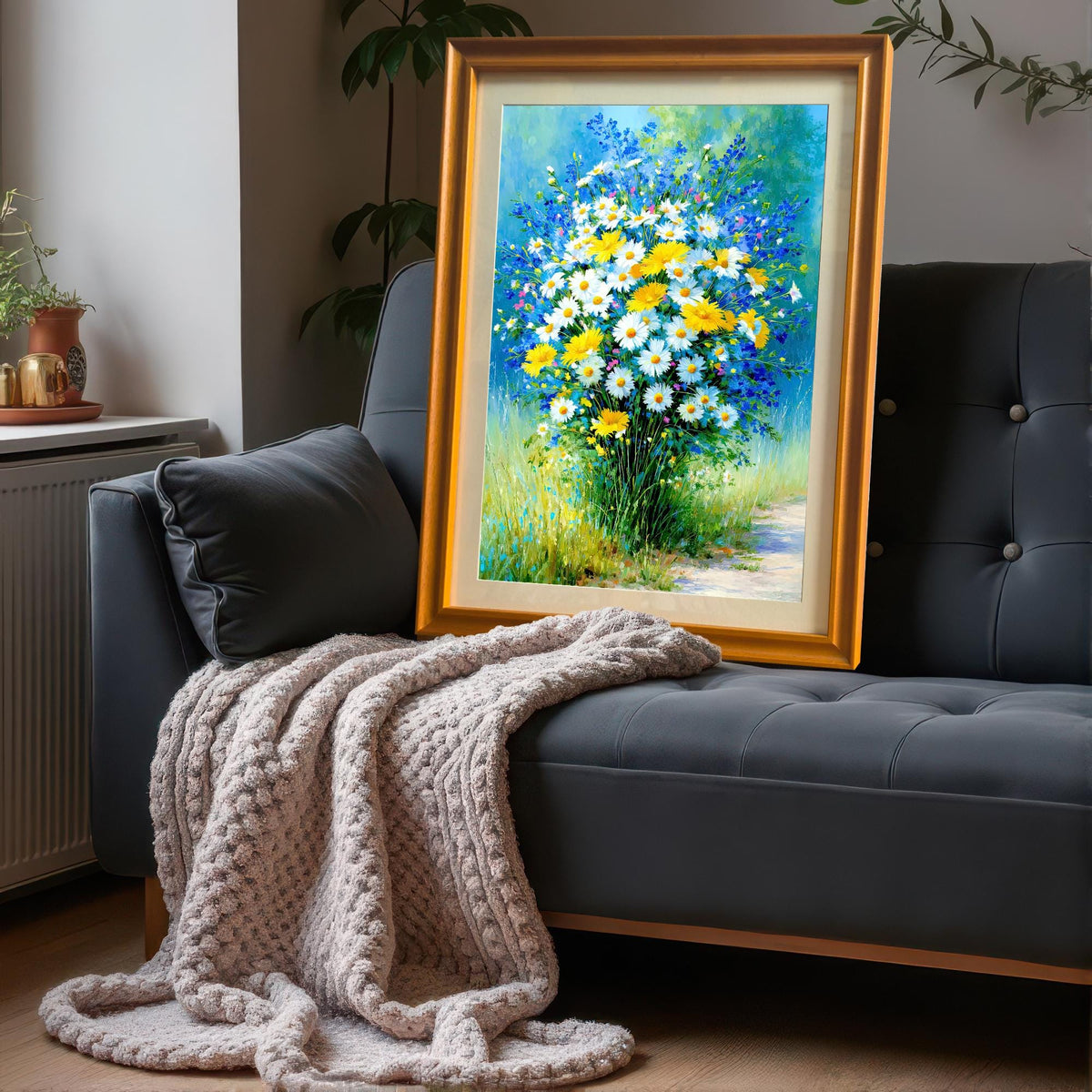 Framed art print with a gold or orange frame leaning on a dark grey sofa with a knitted throw blanket. The art is a colorful impressionist painting of daisies and blue wildflowers.