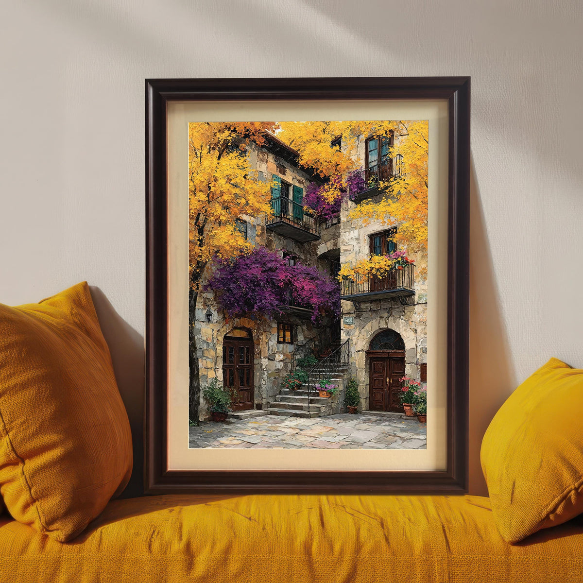 Framed art print with a dark brown wood frame resting on a bright yellow couch between two yellow pillows, showcasing the colorful stone building painting.