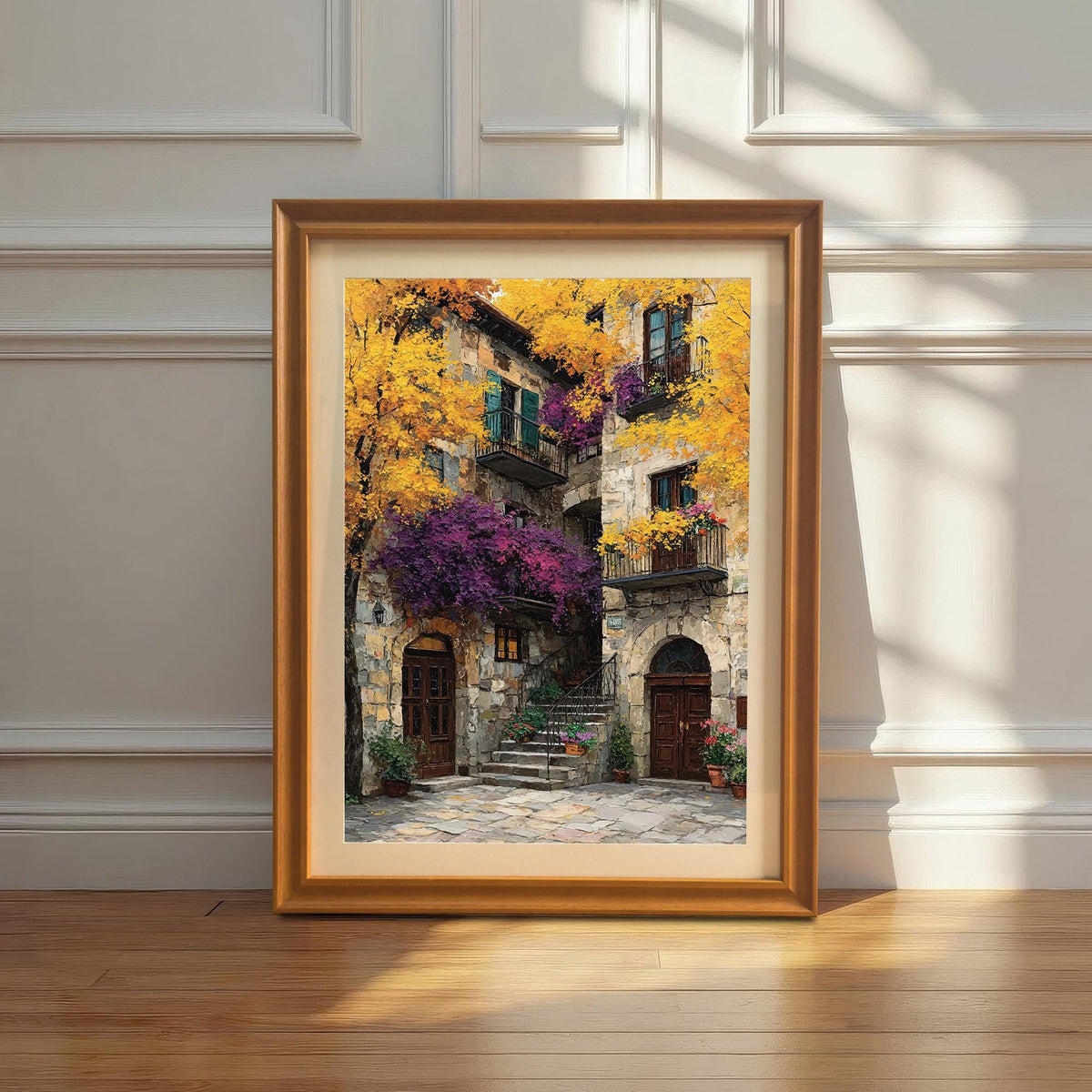 Framed art print with a gold/orange wood frame leaning against a white wainscoted wall. The painting is a vibrant, textured depiction of a Mediterranean stone building covered in bright yellow and purple foliage.
