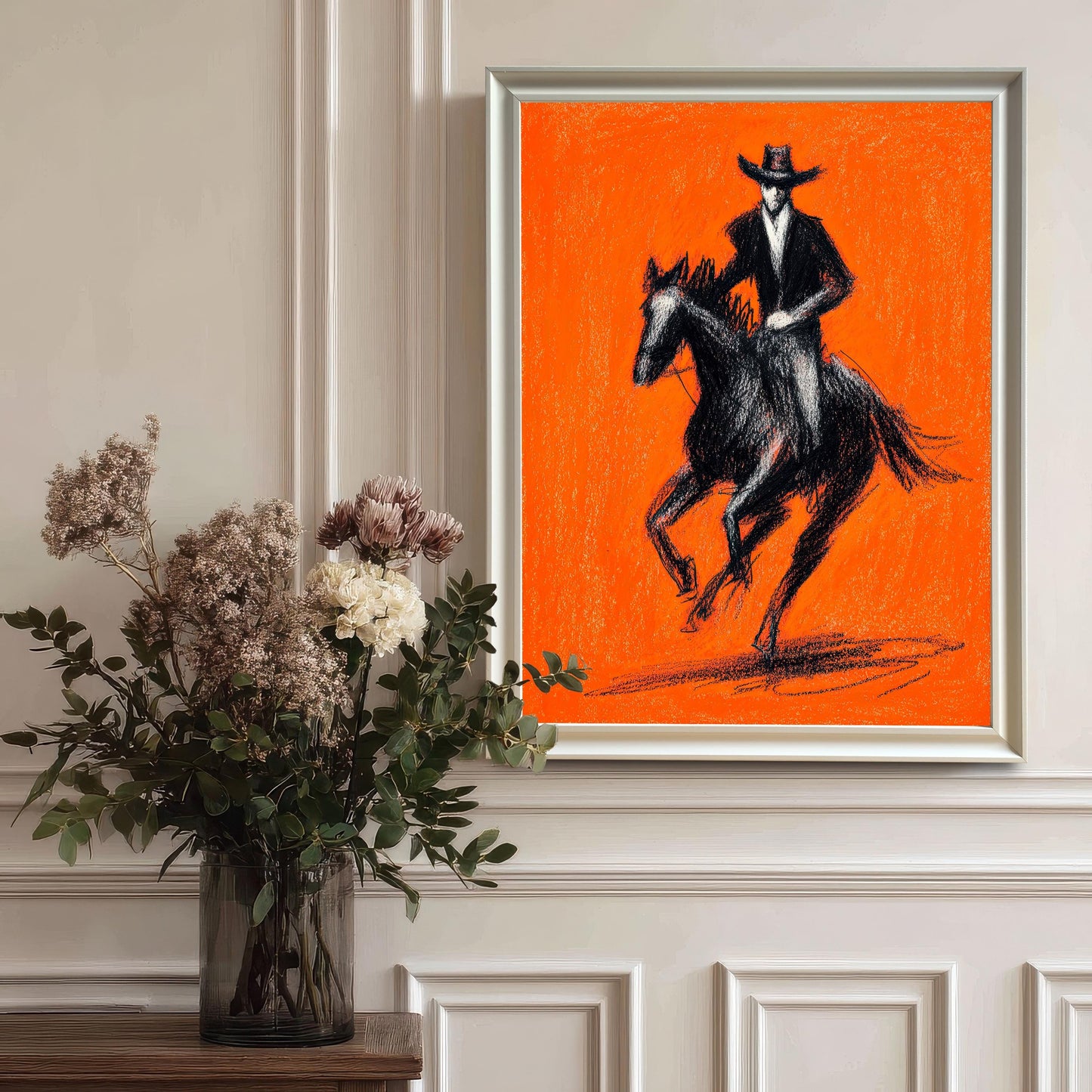 A framed art print of the black and orange cowboy artwork with a White wood frame, hanging on a sophisticated white paneled wall. A vase with dried floral and eucalyptus arrangement sits on a table to the left.