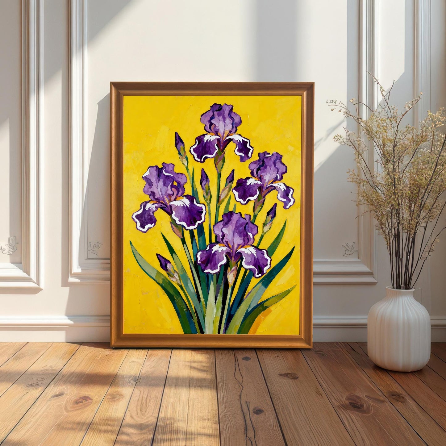A gold-toned Orange framed art print featuring a vibrant painting of purple Irises on a bright yellow background. The framed print is resting on a wood floor against a white paneled wall next to a tall, white vase with dried reeds.